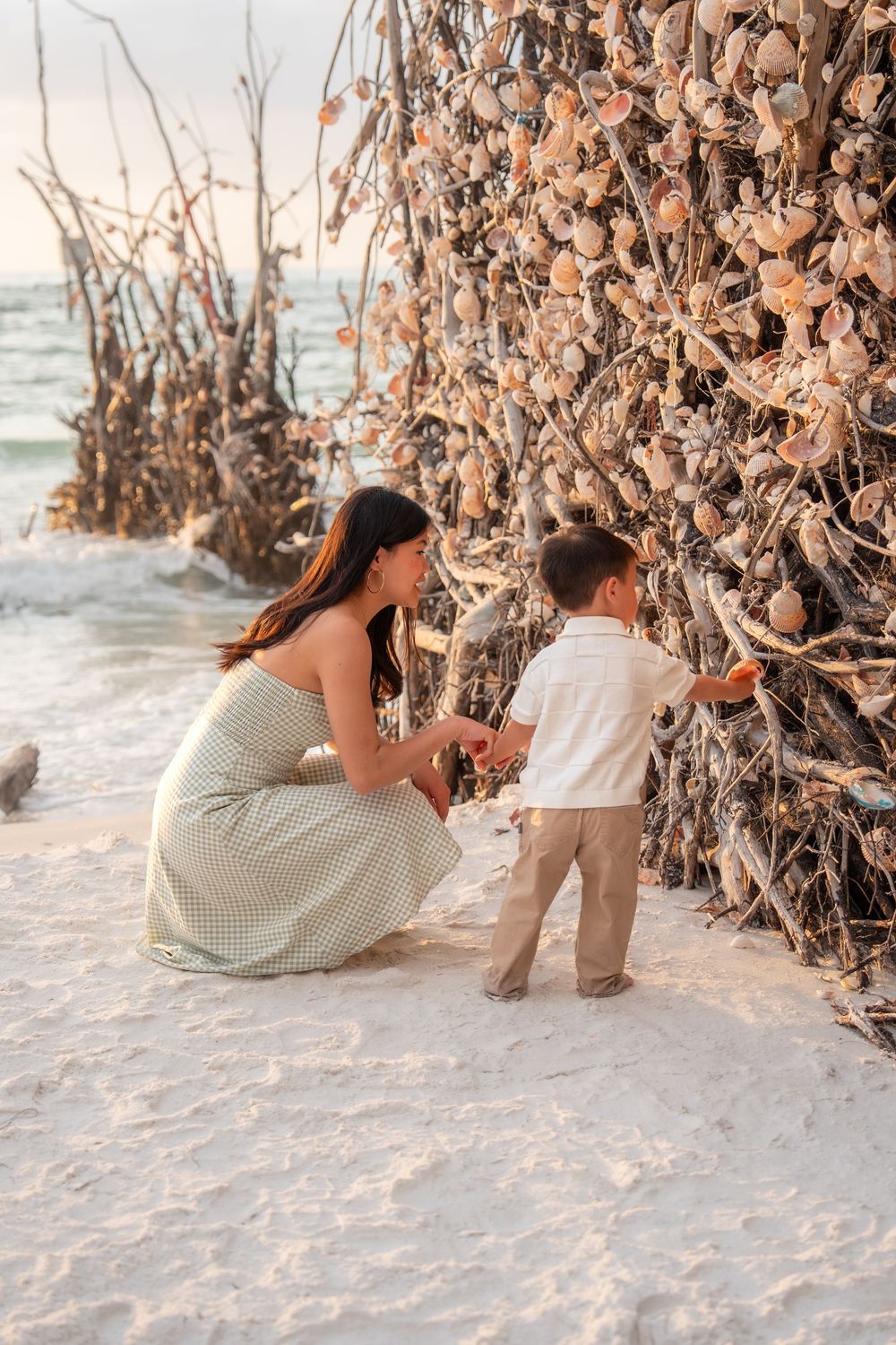 A mother and young son explore a driftwood and seashell art installation on a white sand beach at sunset.