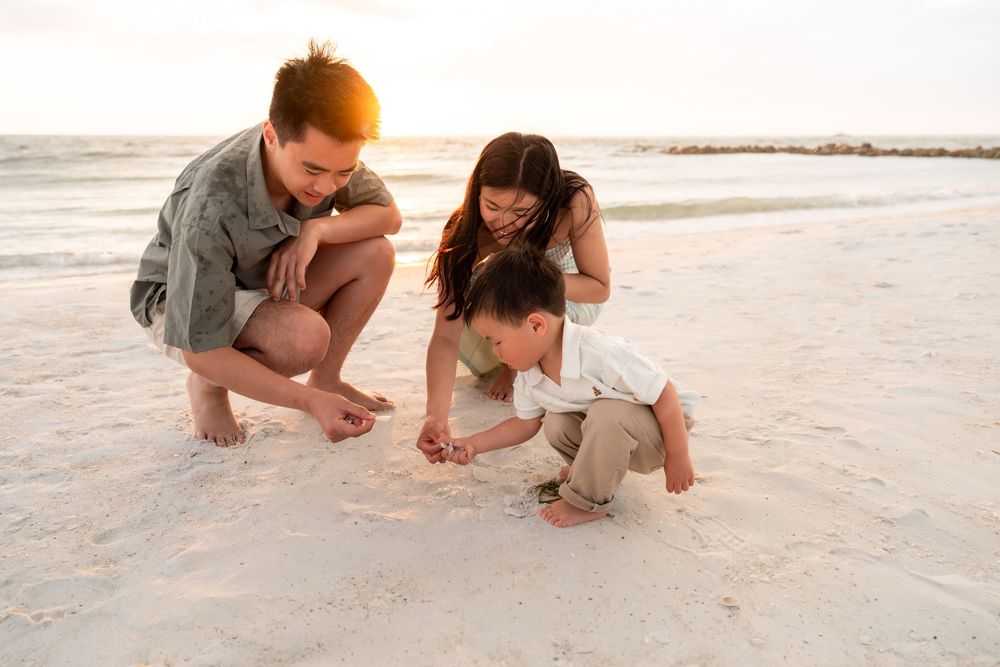 Young family playing in the sand at the beach during golden hour sunset with their toddler son.