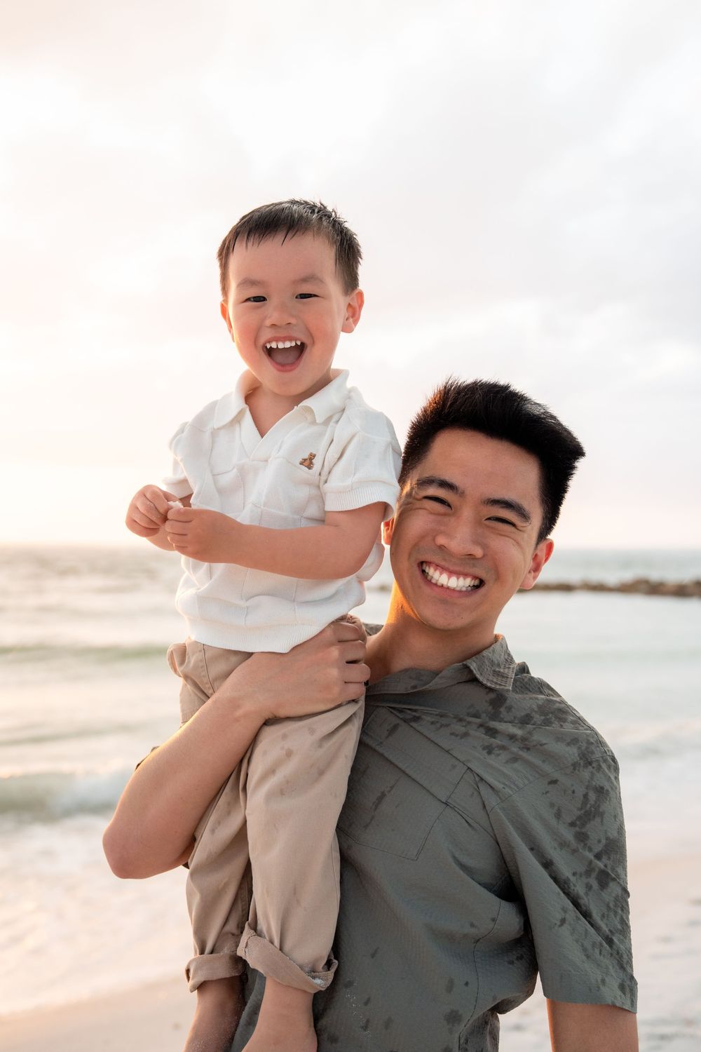 A joyful Asian father carries his laughing toddler son on his shoulders at a beach during sunset.