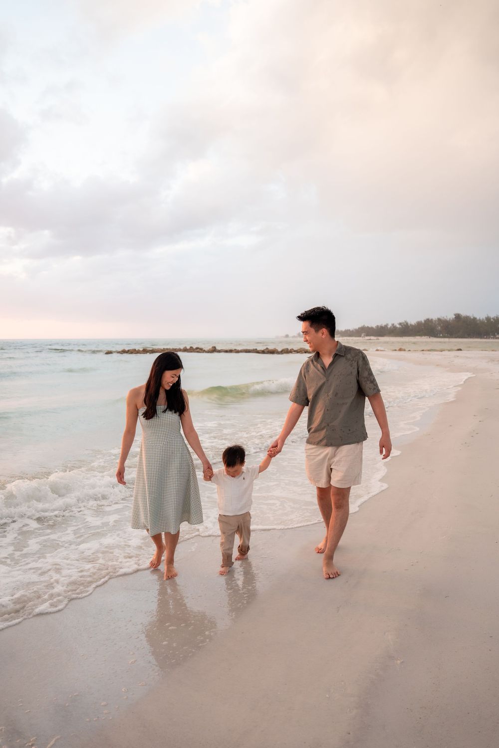 Happy Asian family walks along sandy beach at sunset, parents holding hands with their toddler son.