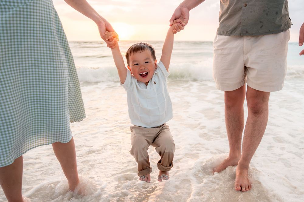 Happy toddler boy being swung by parents on a sunny beach at sunset, waves lapping at their feet.
