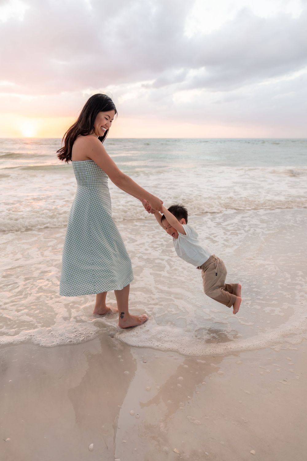 A joyful mother swings her young son by the hands along a beautiful sunset beach shoreline.