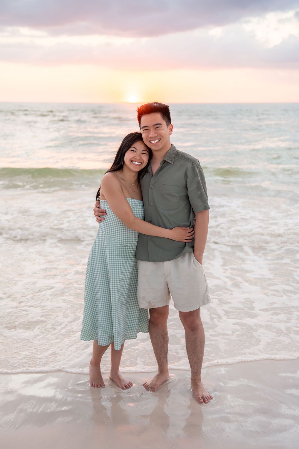 A smiling Asian couple embraces on a sandy beach during a vibrant ocean sunset.