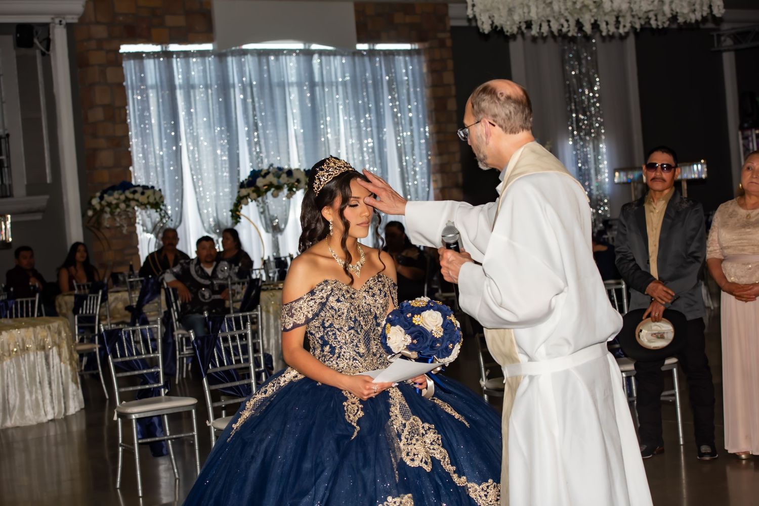 A quinceañera girl in a navy blue and gold gown receives her crown from a priest at an elegant reception hall.