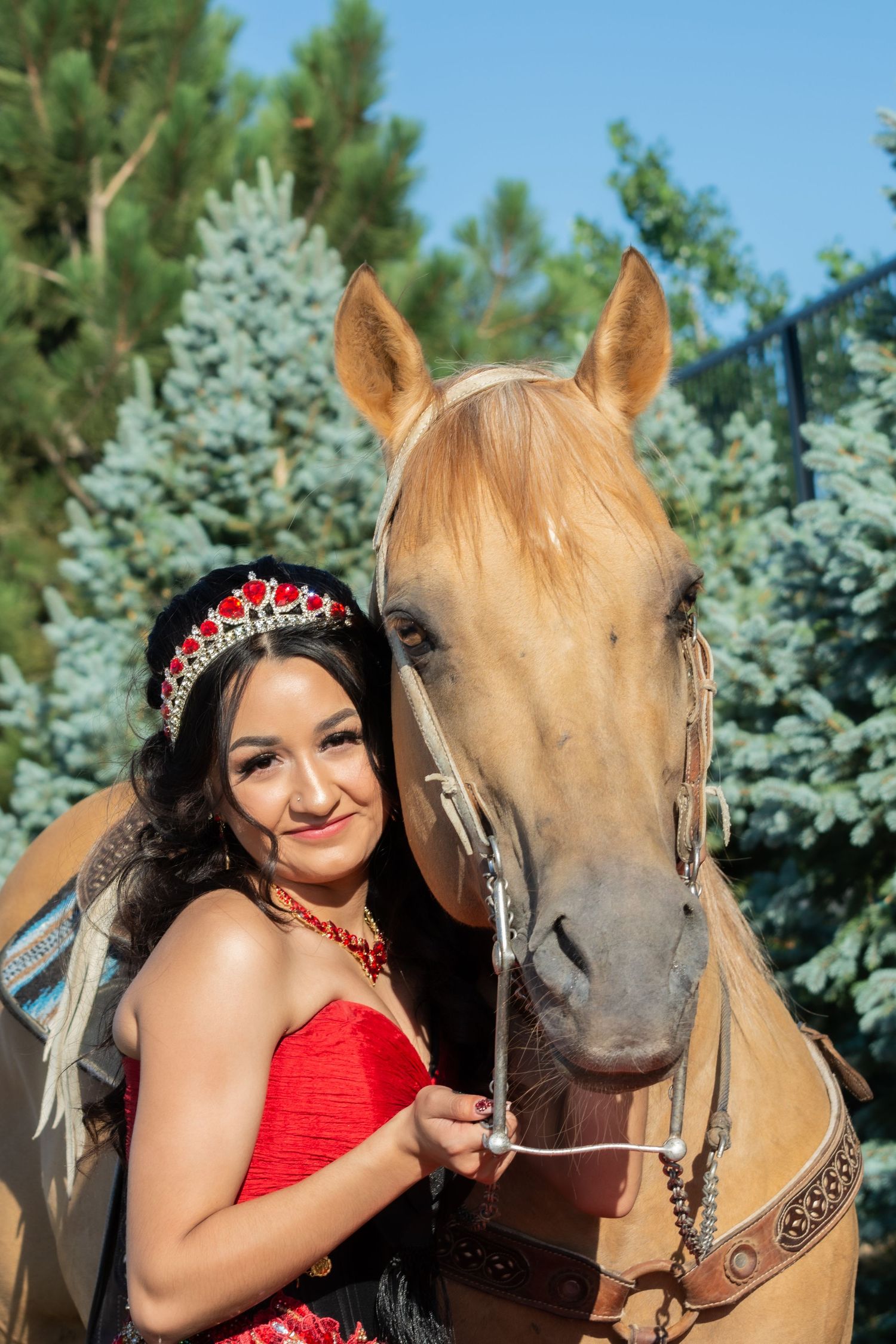 Smiling young woman in red quinceañera dress with tiara stands beside palomino horse among pine trees.