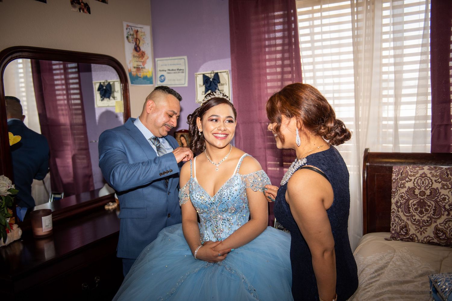A young woman in a light blue quinceañera dress is helped by her parents while getting ready at home.