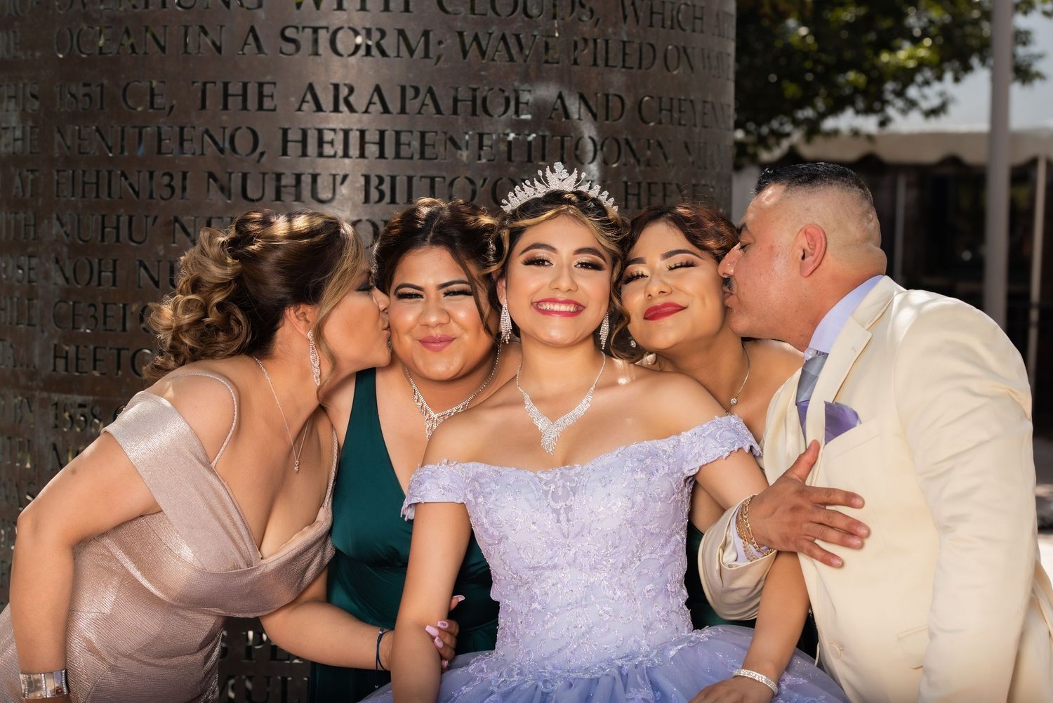 Quinceañera girl in lavender gown and tiara receives kisses from family members at outdoor celebration.