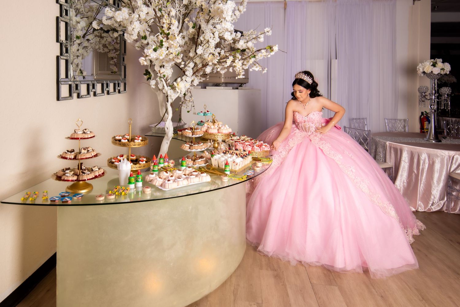 Quinceañera girl in pink ball gown leans over elegant dessert table with cupcakes and sweet treats.