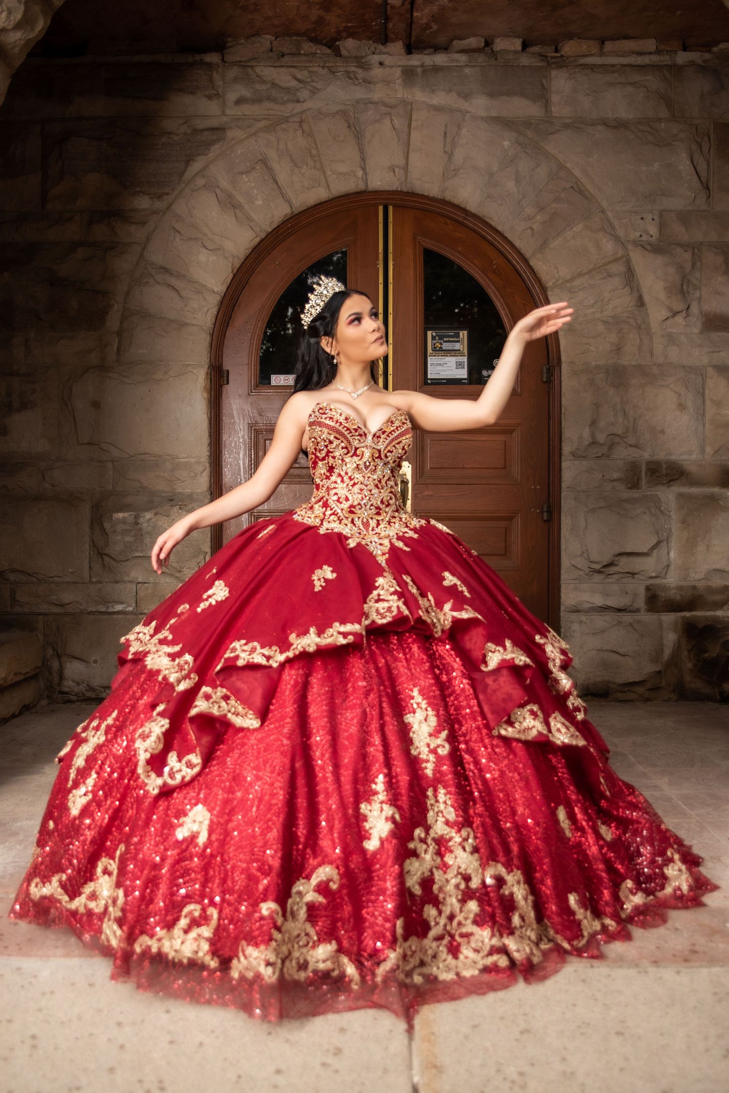 Young woman in red and gold embroidered quinceañera ball gown poses gracefully before stone archway.