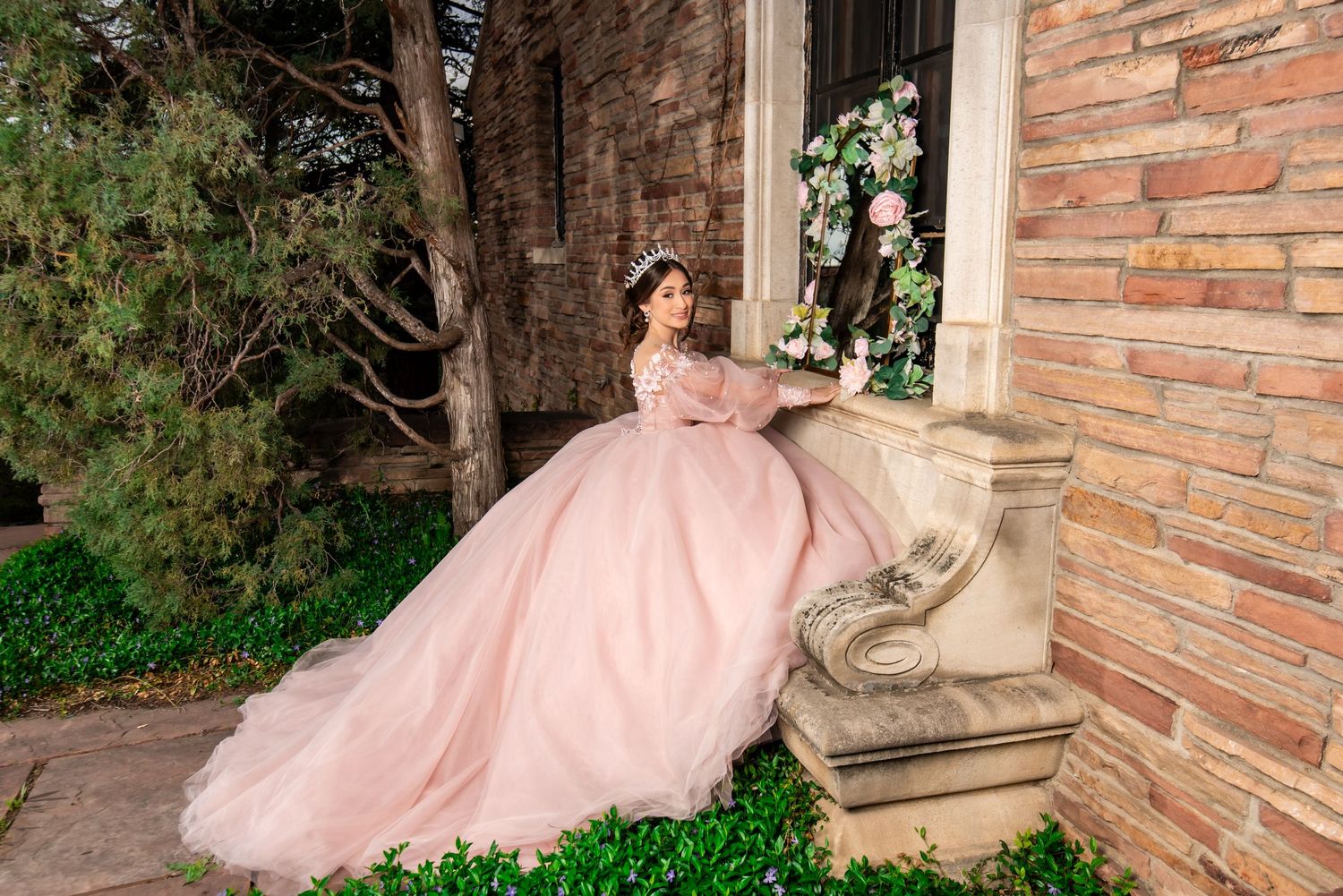 Young woman in pink ball gown quinceañera dress poses elegantly beside brick wall with floral decorations.