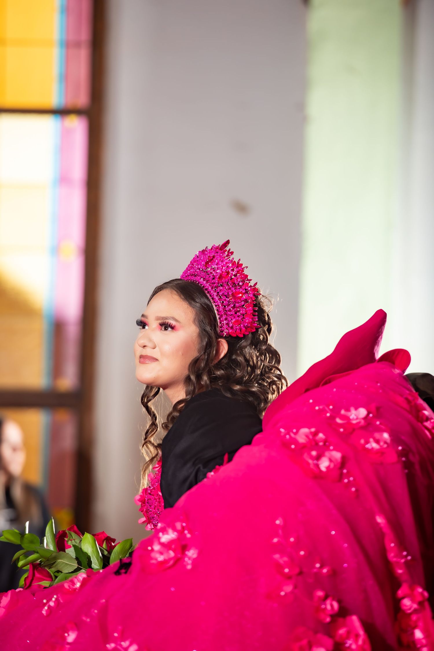 Quinceañera girl in vibrant pink gown and matching crown gazes upward holding roses at celebration.