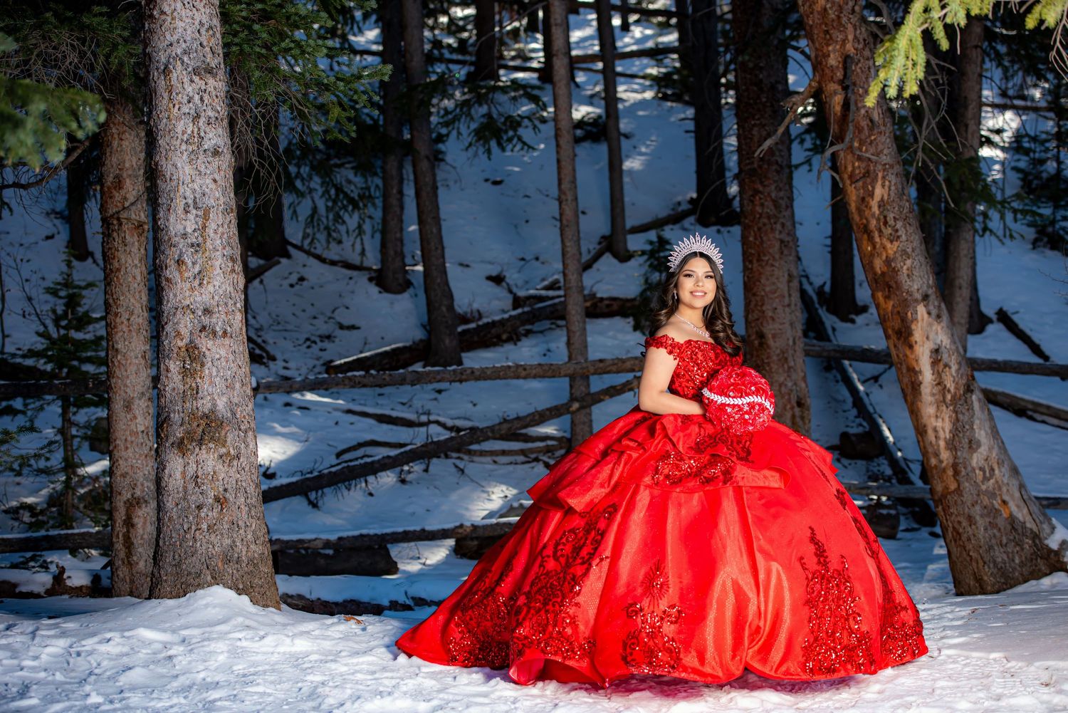 Young woman in red quinceañera ball gown with embroidery poses among snow-covered trees in winter forest.