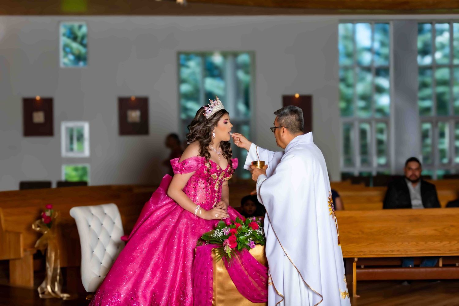 A quinceañera in a pink ball gown receives a blessing from a priest during a church ceremony.