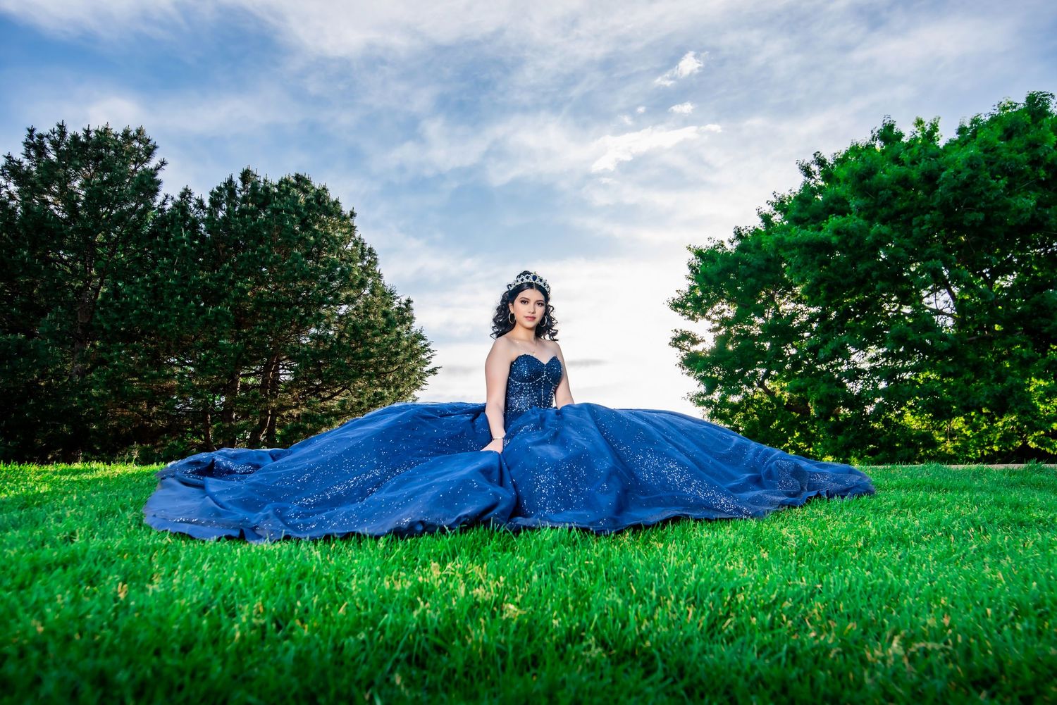 Quinceañera girl in royal blue ball gown with tiara sits gracefully on green grass under cloudy sky.