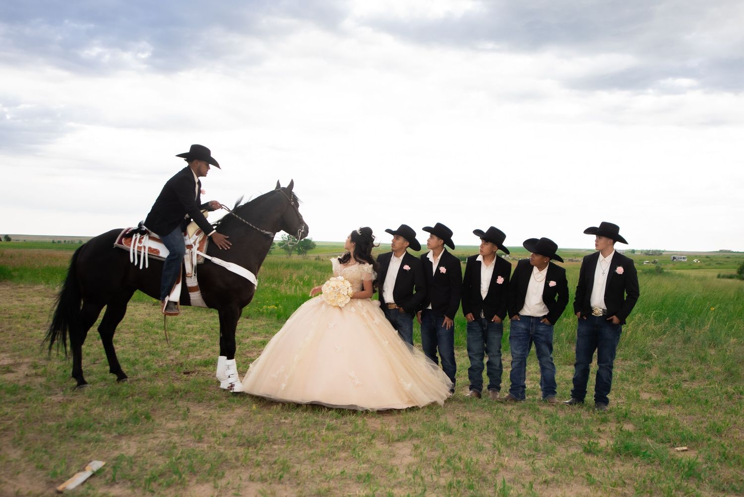 Quinceañera girl in champagne gown poses with chambelanes in cowboy hats beside black horse in field.