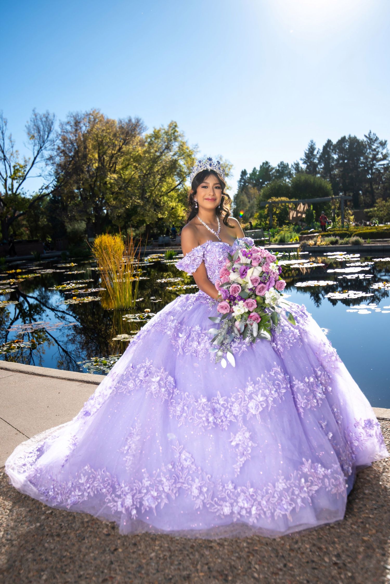 Quinceañera girl in lavender floral ball gown holds bouquet beside serene lily pond in sunny garden.