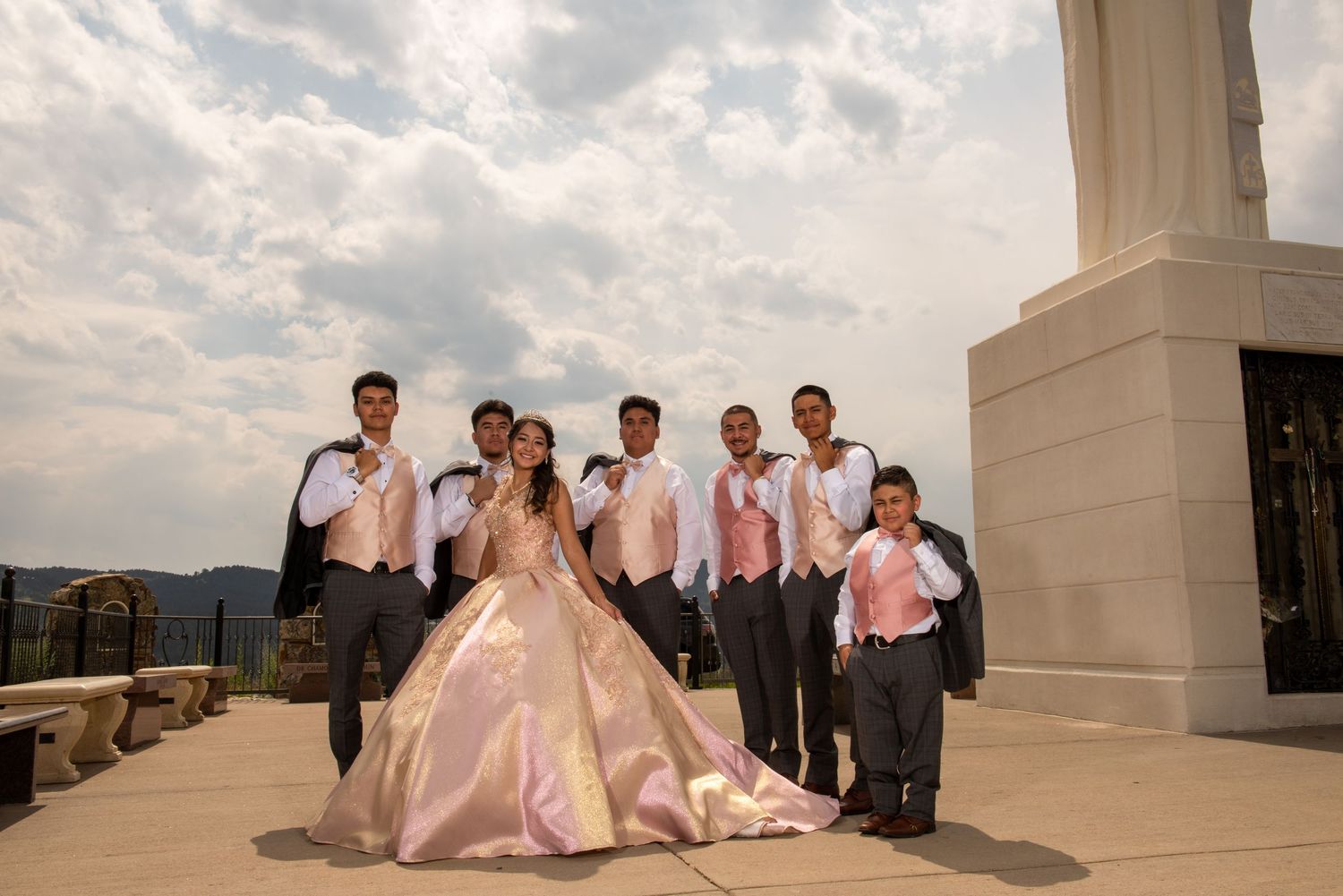 A quinceañera in a blush pink gown poses with her chambelanes in gray suits outdoors under cloudy skies.