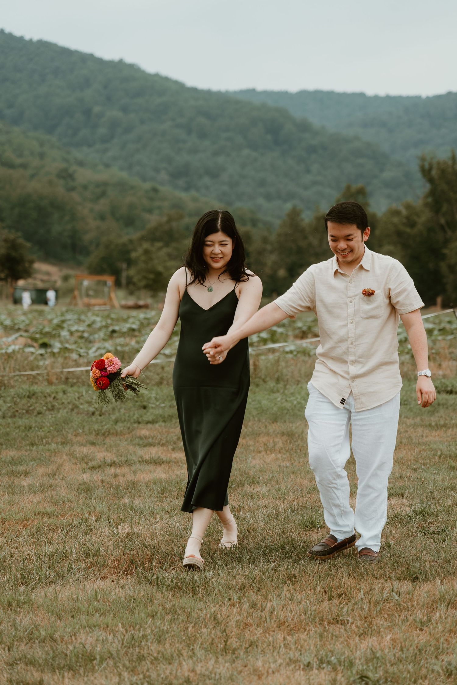 Couple holding hands walking through green field with mountain backdrop, woman in black dress holding red bouquet.