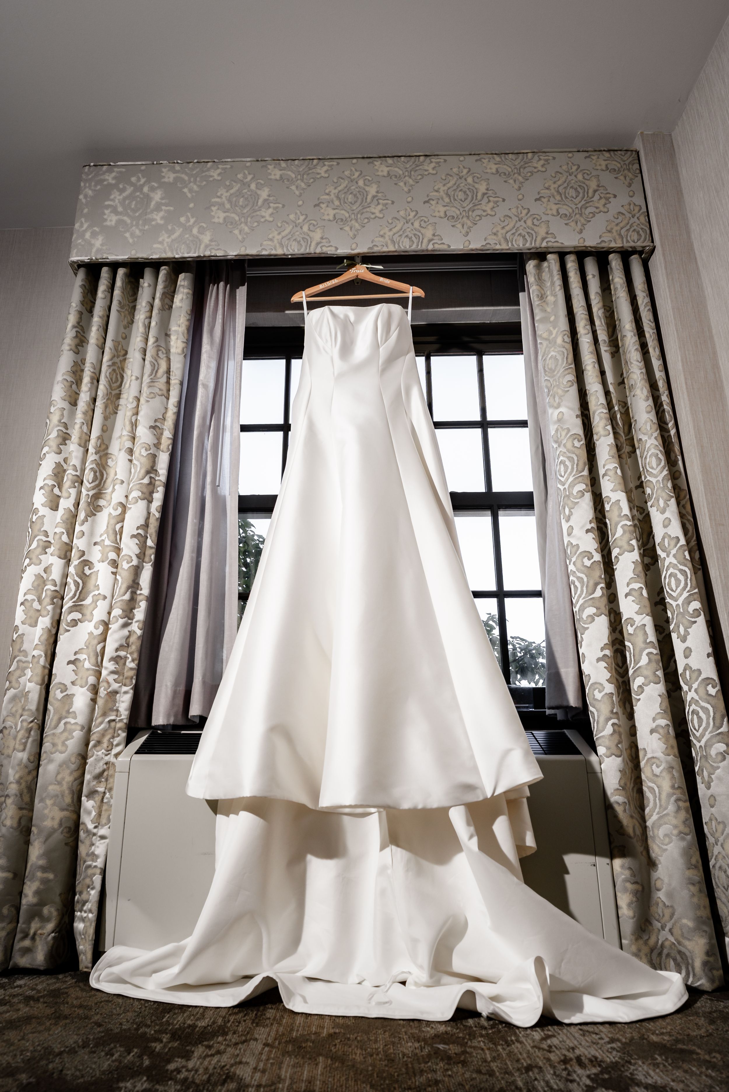 Elegant white strapless wedding gown with a long train hanging on a wooden hanger in front of a draped window.