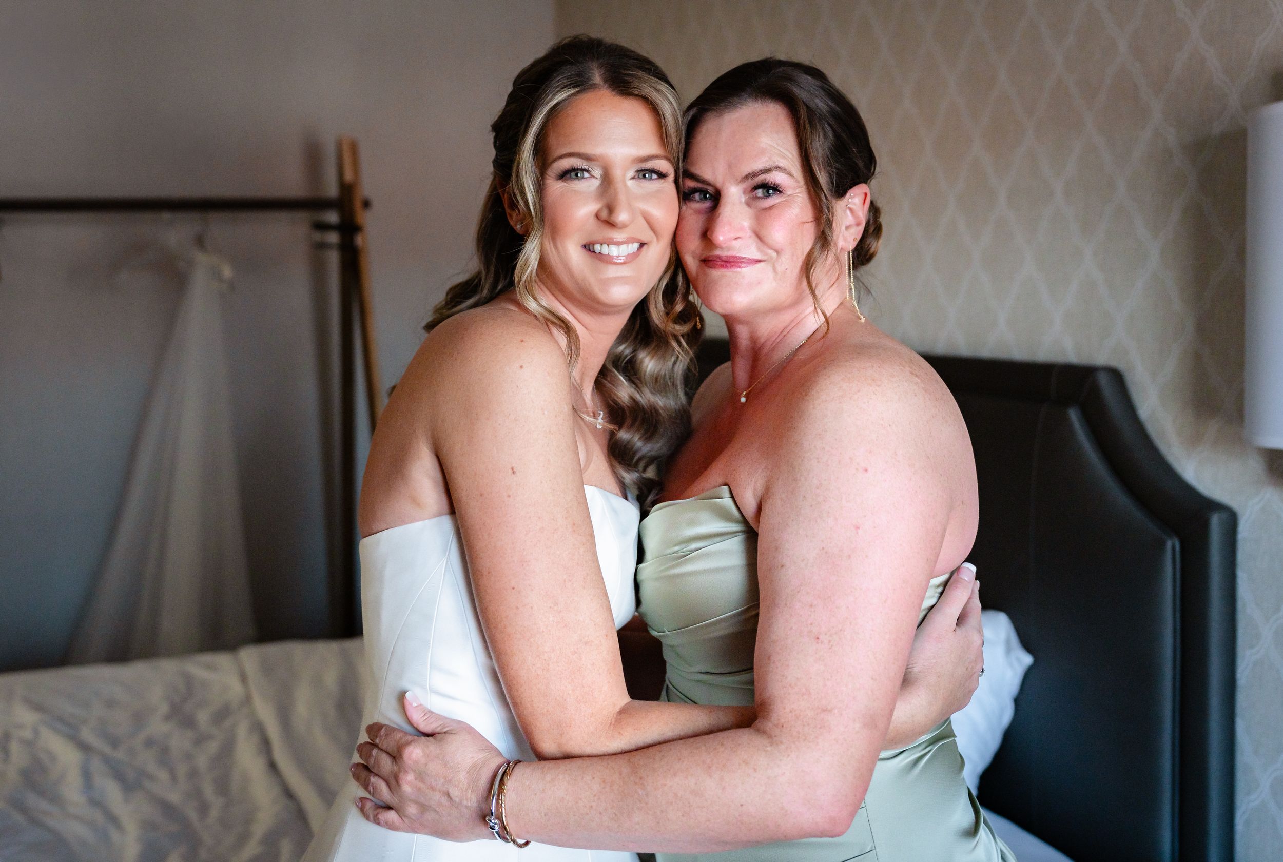 Bride and bridesmaid sharing a warm embrace in sage green dress during wedding preparations in hotel room.