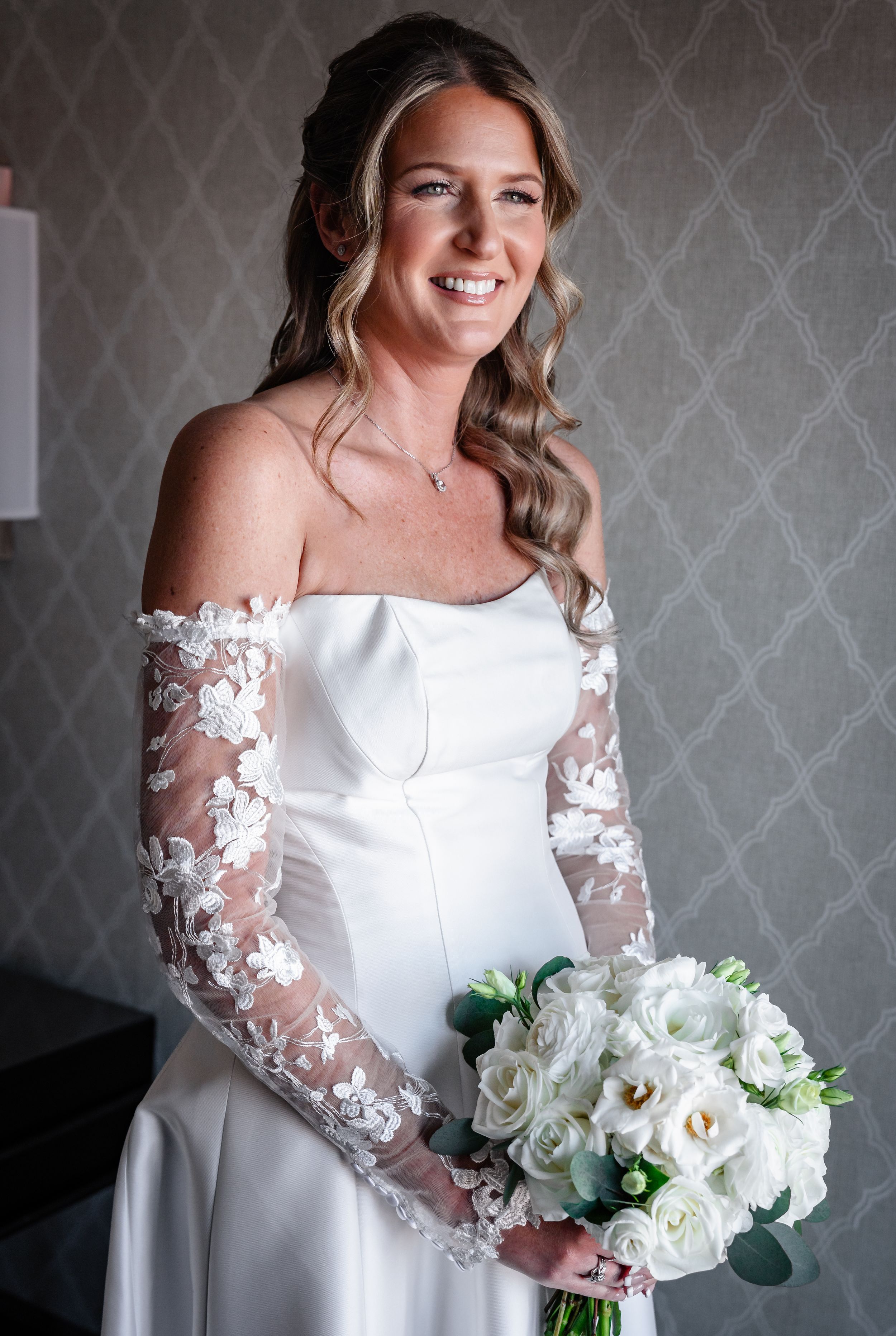 Smiling bride in off-shoulder lace wedding gown holding white rose bouquet on wedding day.