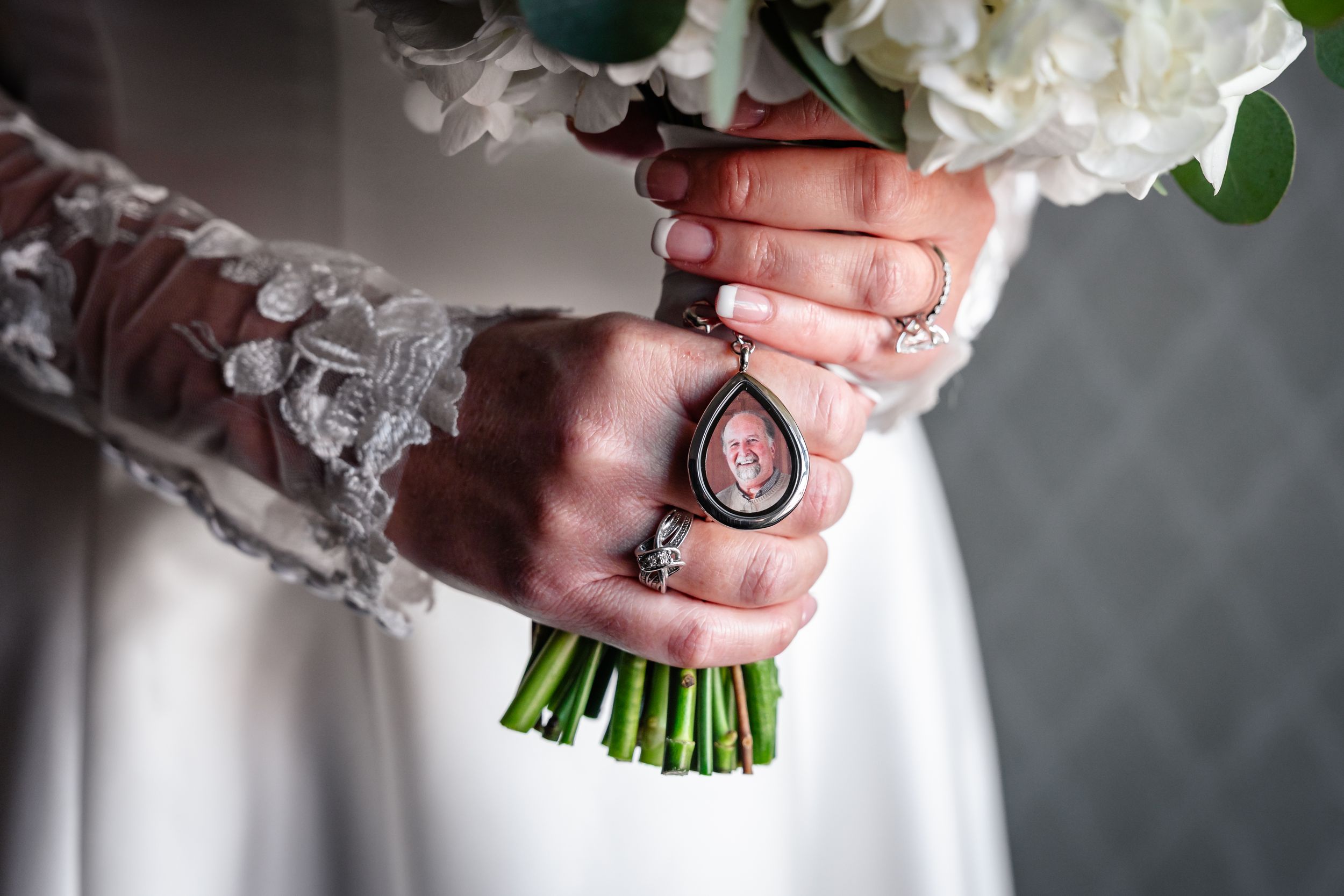 Close-up of bride holding white peony bouquet with a memorial photo charm pendant on wedding day.