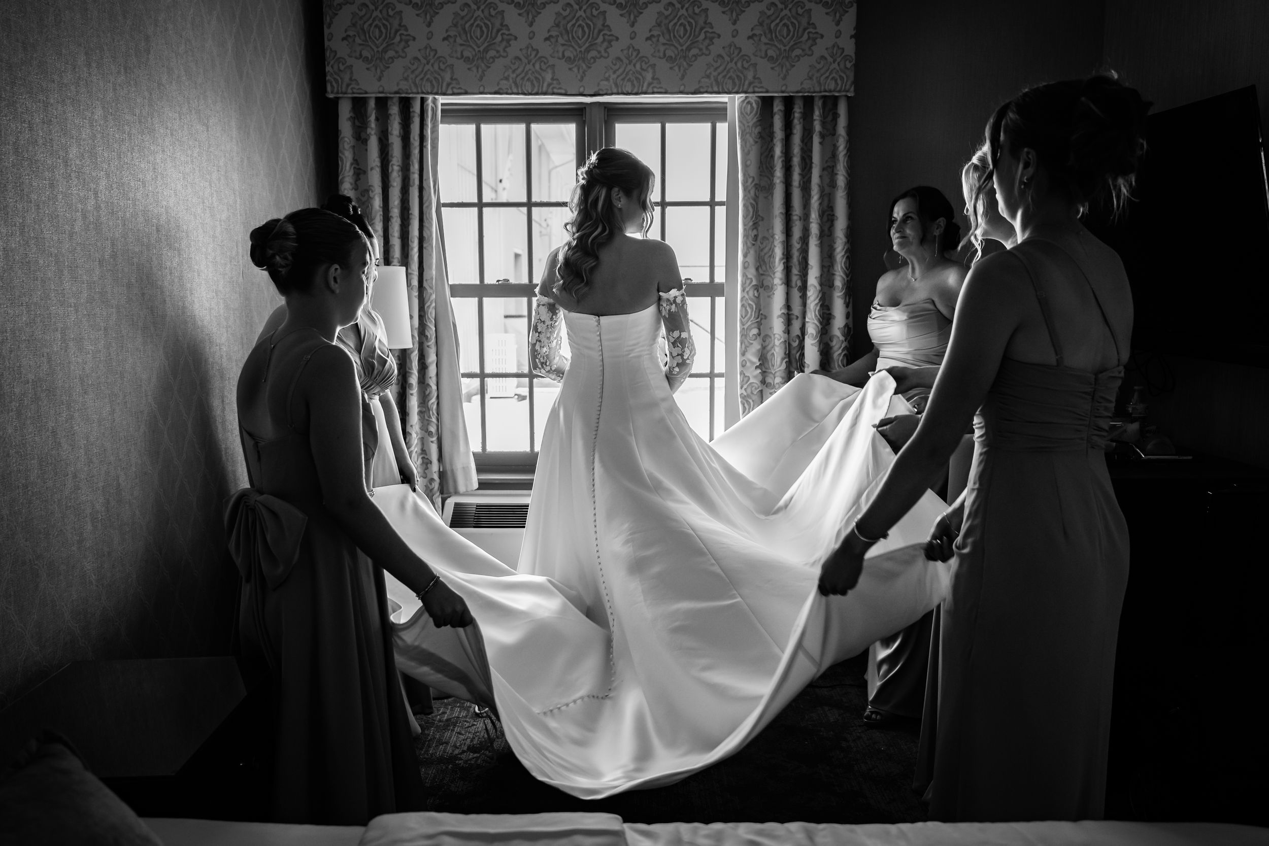 Black and white photo of bridesmaids helping bride with wedding dress train by window.