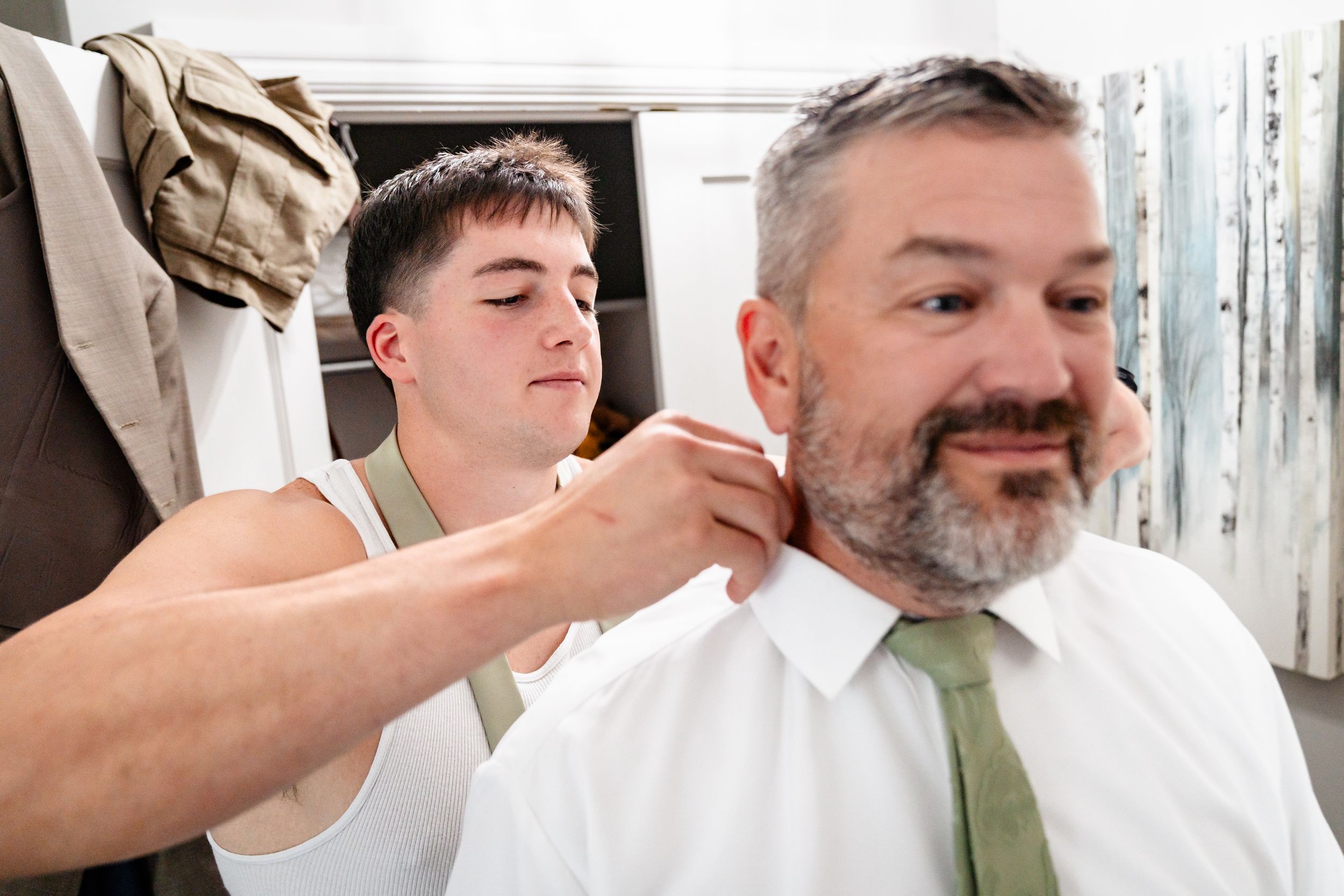 Young groomsman carefully adjusting grooms tie during wedding day preparations indoors.