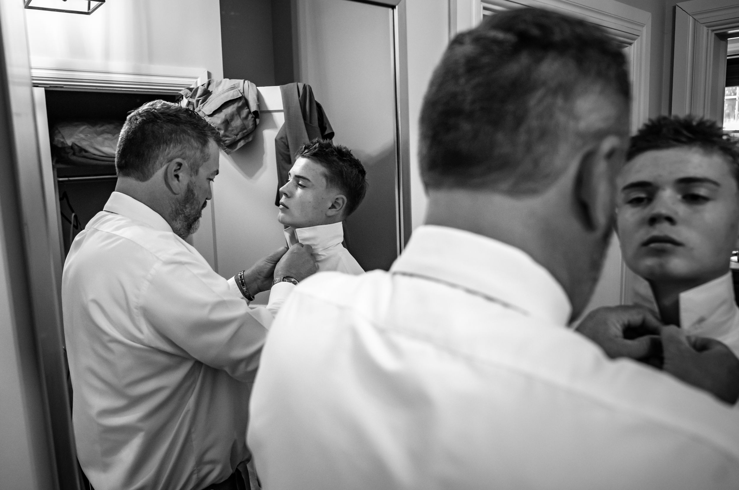 Black and white photo of groomsmen helping each other with ties in mirror before wedding.
