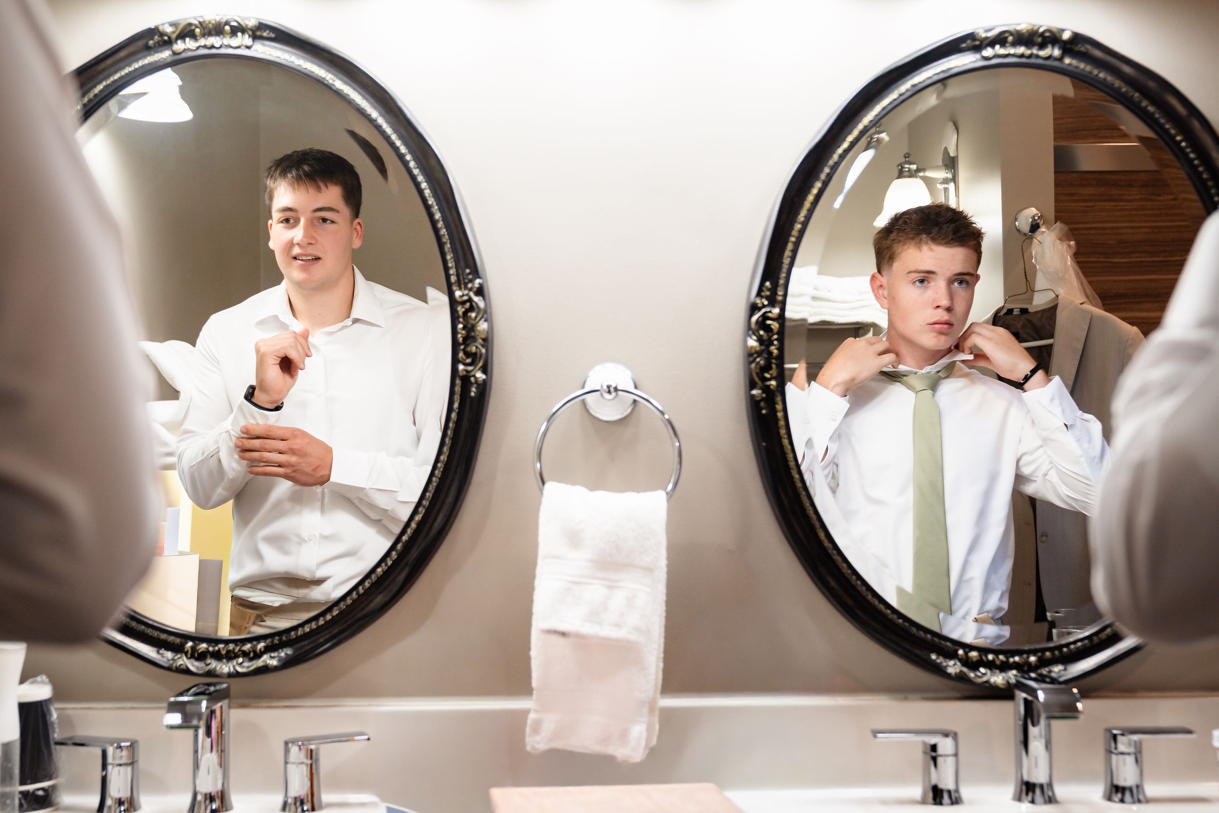 Young groomsman adjusting his green tie while looking in ornate oval bathroom mirror before wedding.