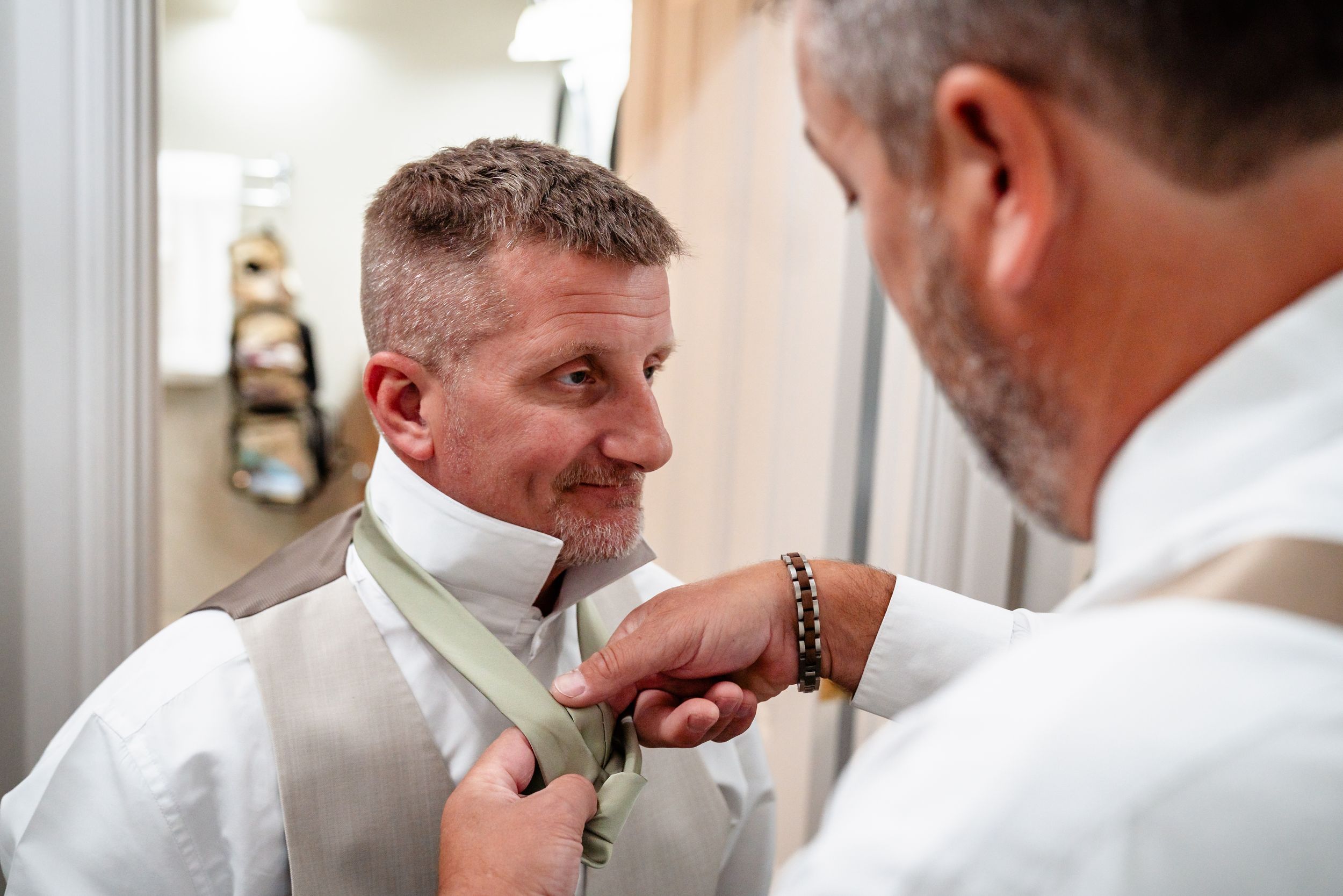 Groomsman helping groom adjust tie during wedding day preparations.