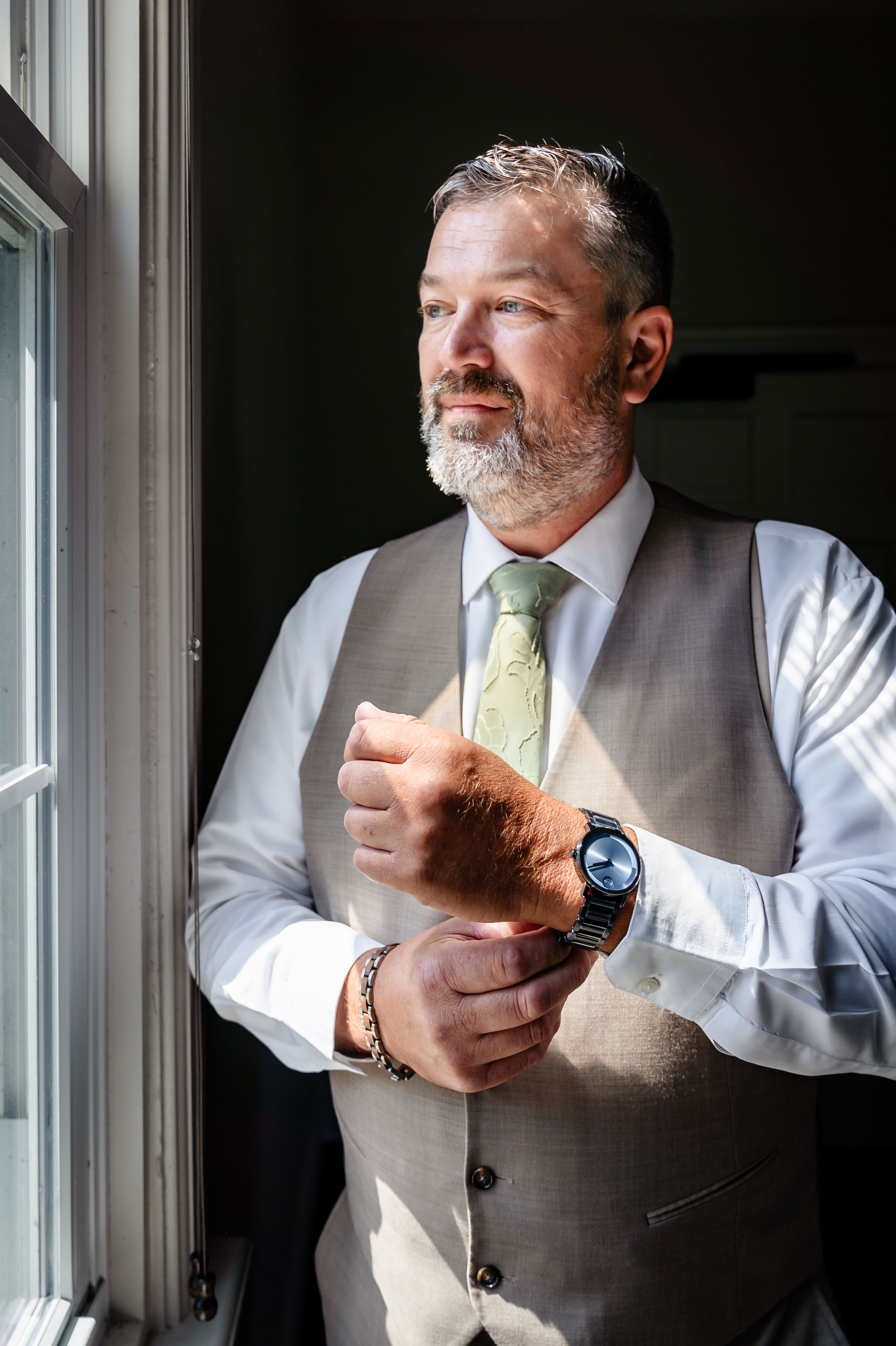 Groom in gray vest and green tie adjusting cufflinks by sunlit window.