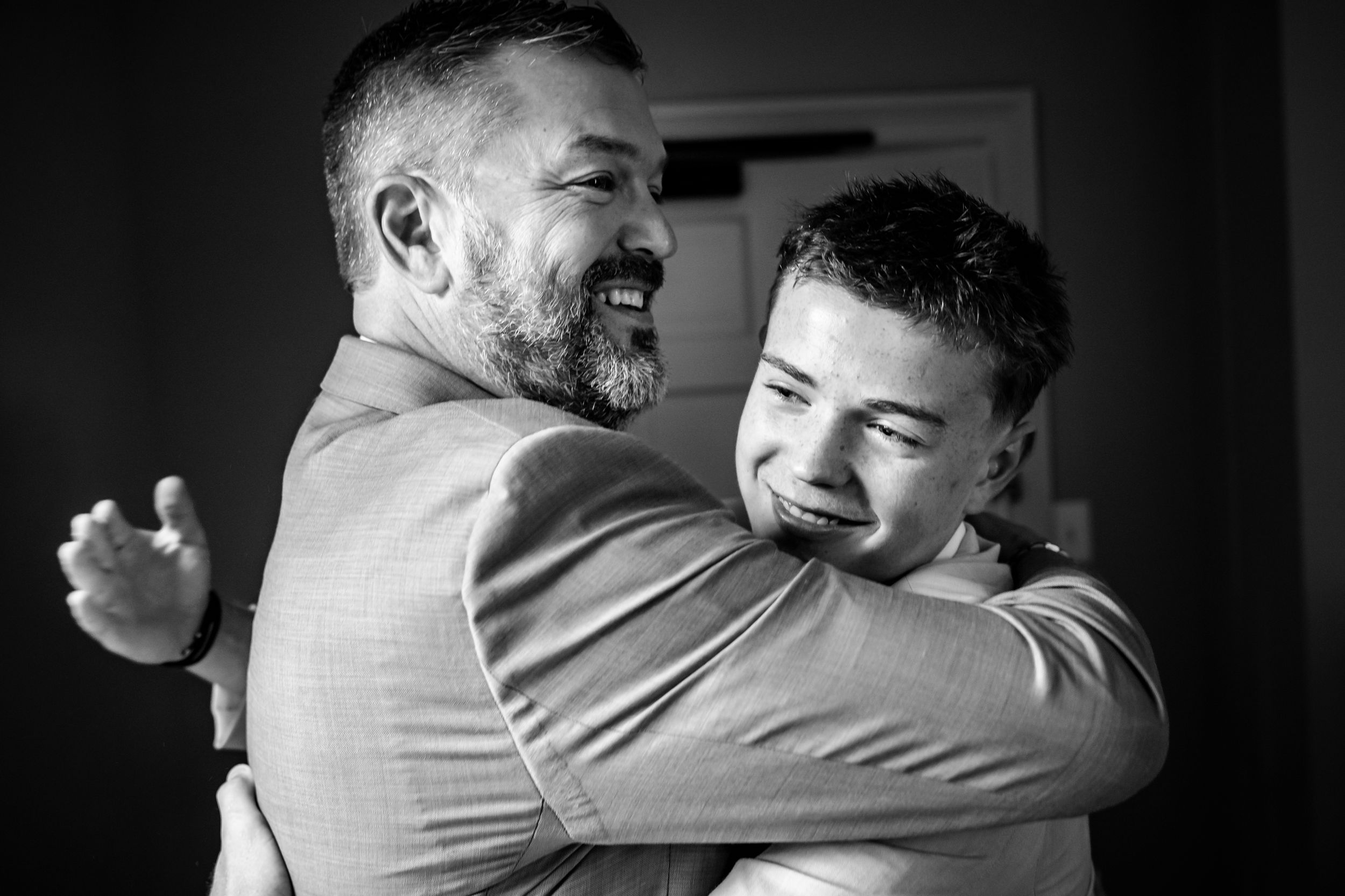 Groom sharing joyful hug with young boy in black and white wedding photo.