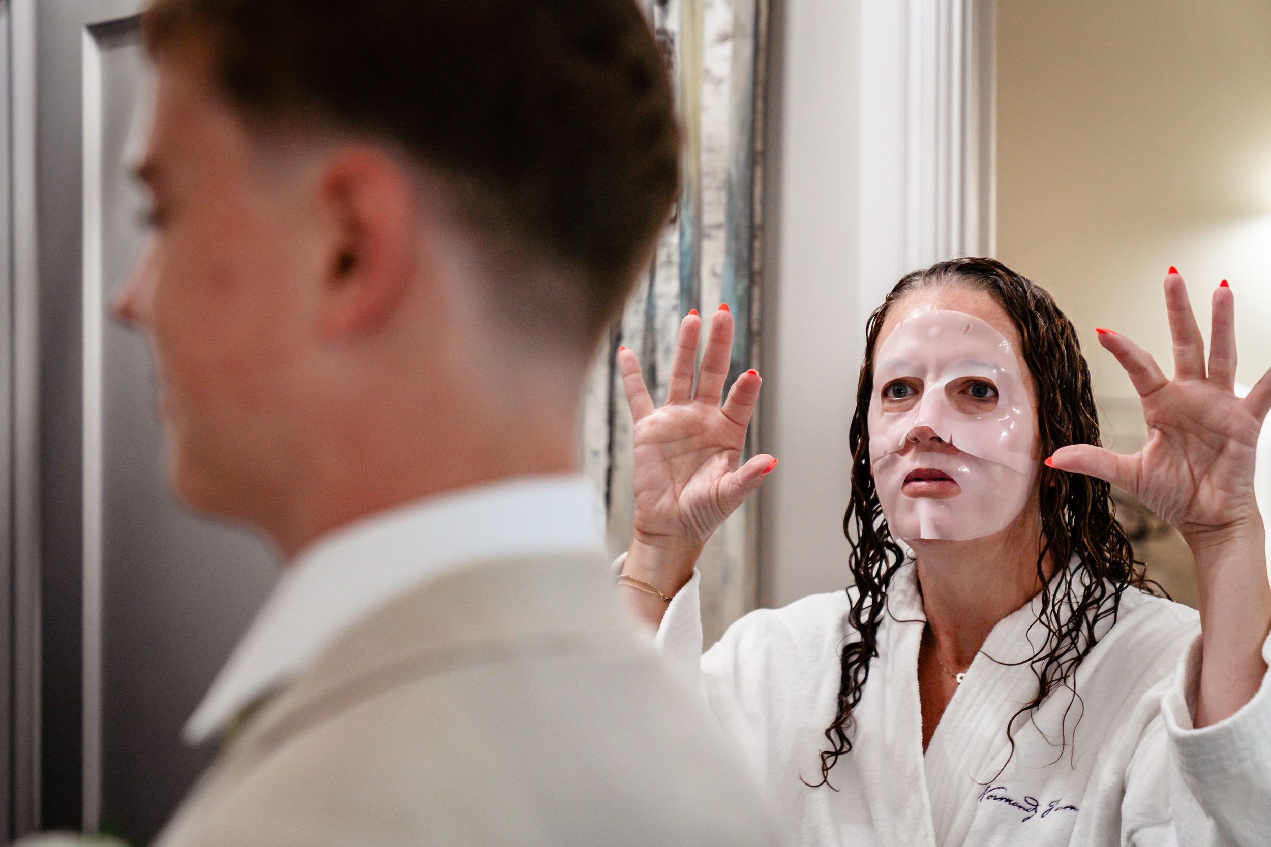 Bride wearing facial sheet mask posing dramatically in mirror before wedding.