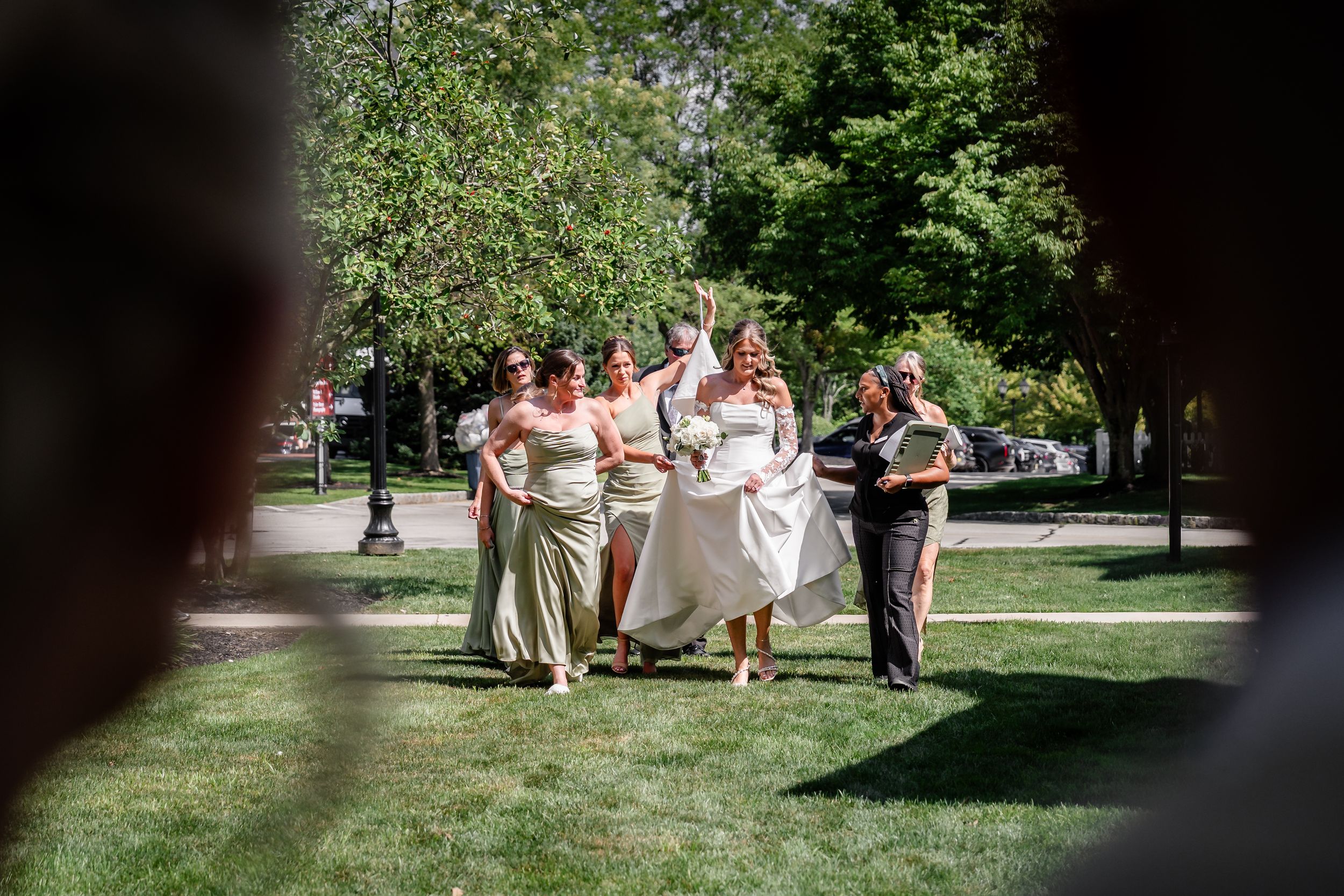 Wedding party walks across green park lawn with bride in white gown and bridesmaids in gold.
