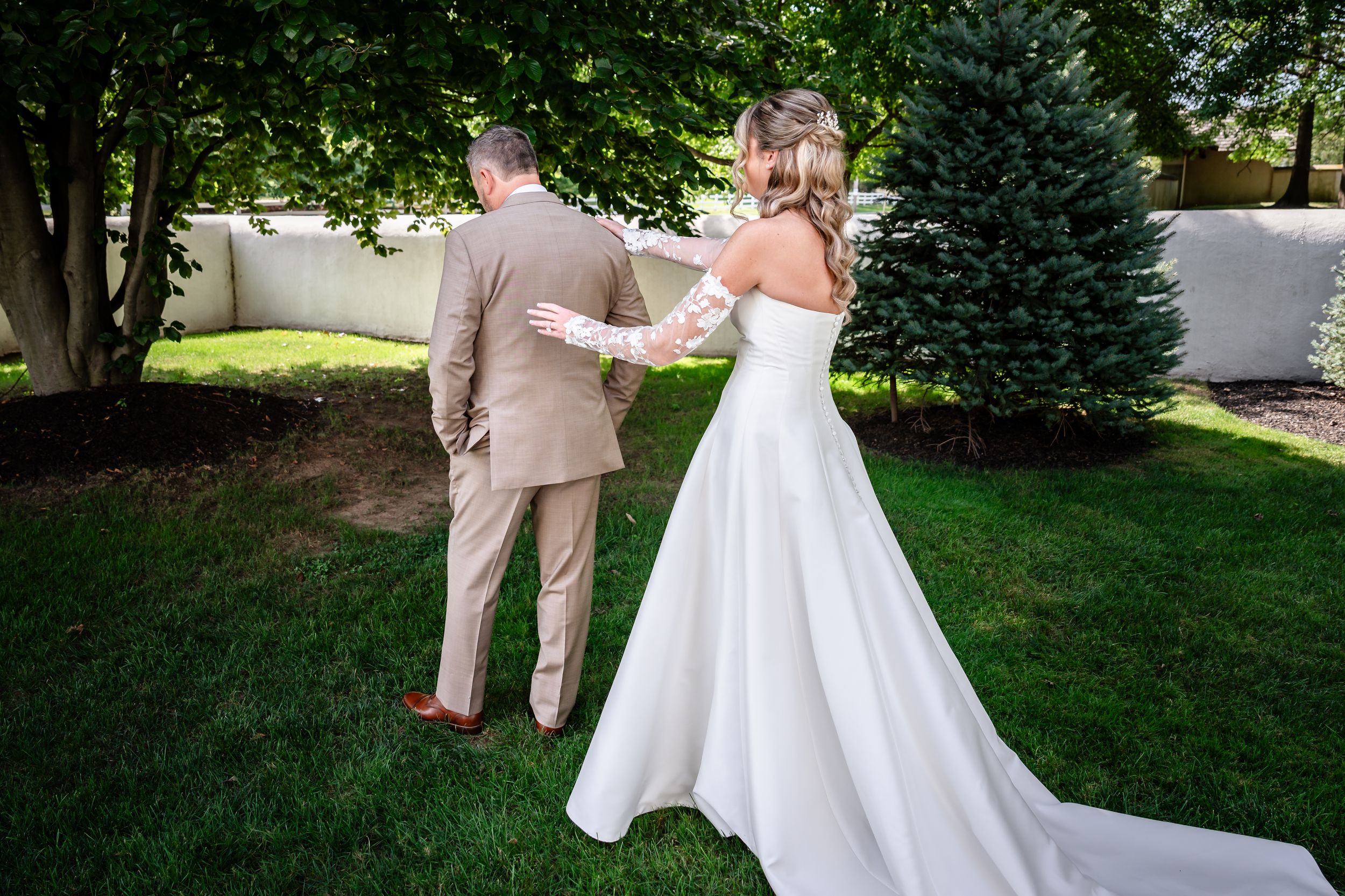 Bride in white off-shoulder gown tapping groom's shoulder during outdoor first look moment.