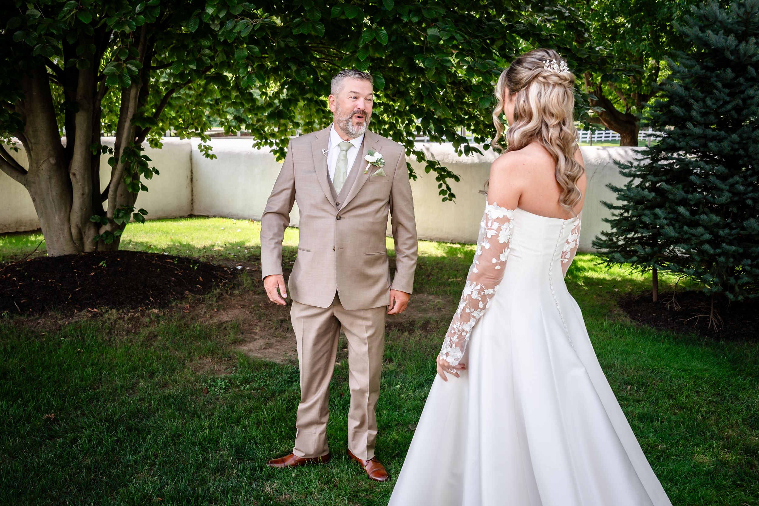 Groom in tan suit turns to see bride in lace wedding dress during outdoor first look.