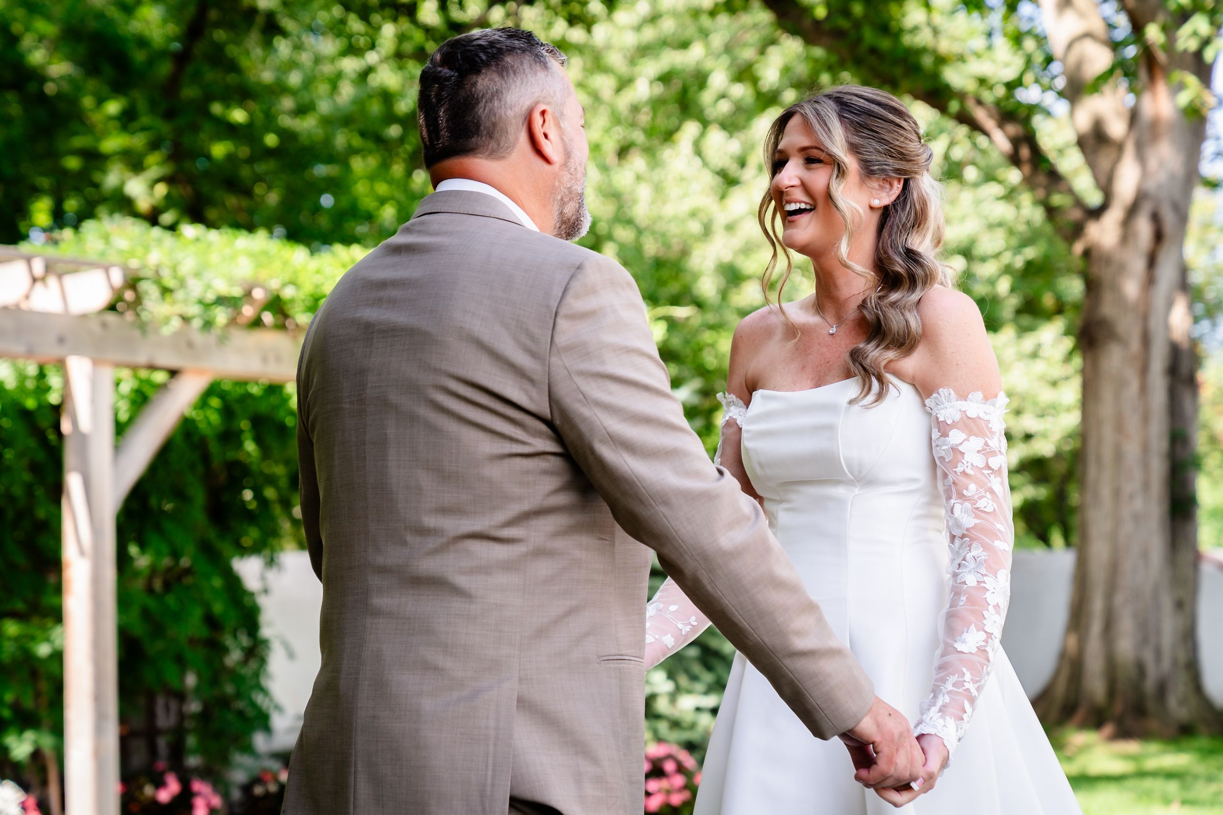 Bride and groom share joyful first look moment holding hands in sunny garden setting.