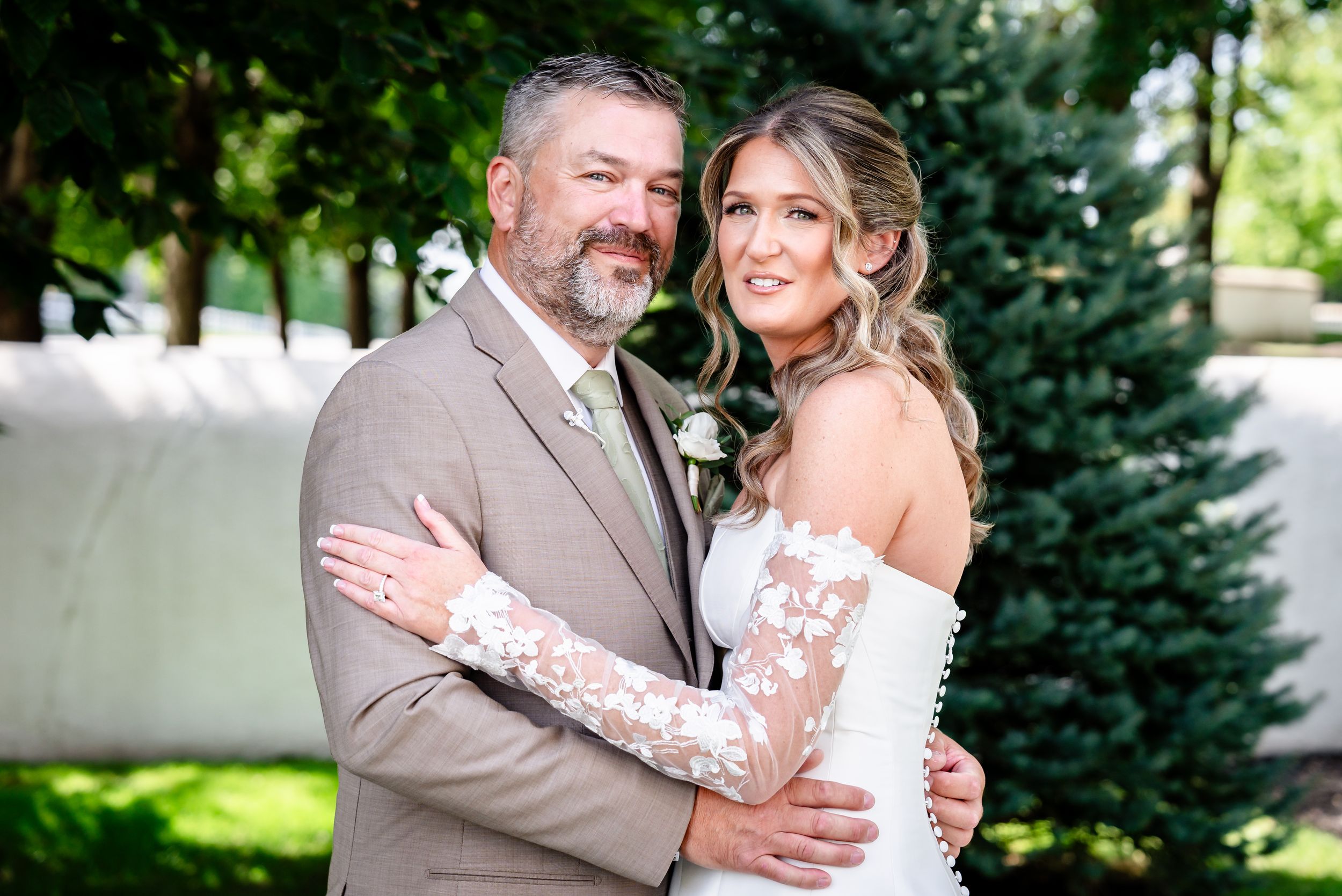 Bride and groom pose together outdoors, bride wearing lace sleeve wedding gown, groom in tan suit.