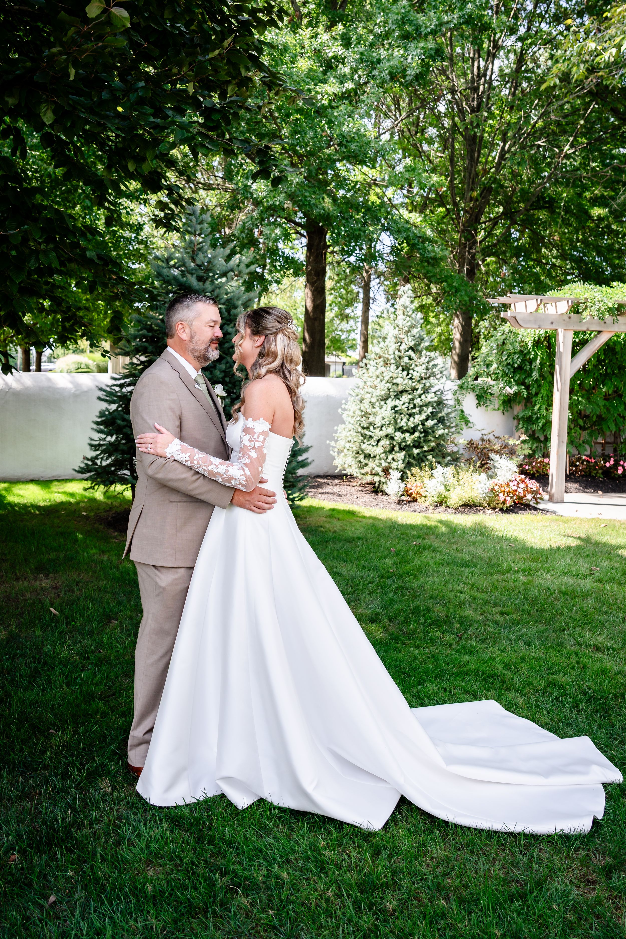 Bride and groom share a romantic embrace in a lush garden, bride wearing a stunning white gown with floral lace sleeves.