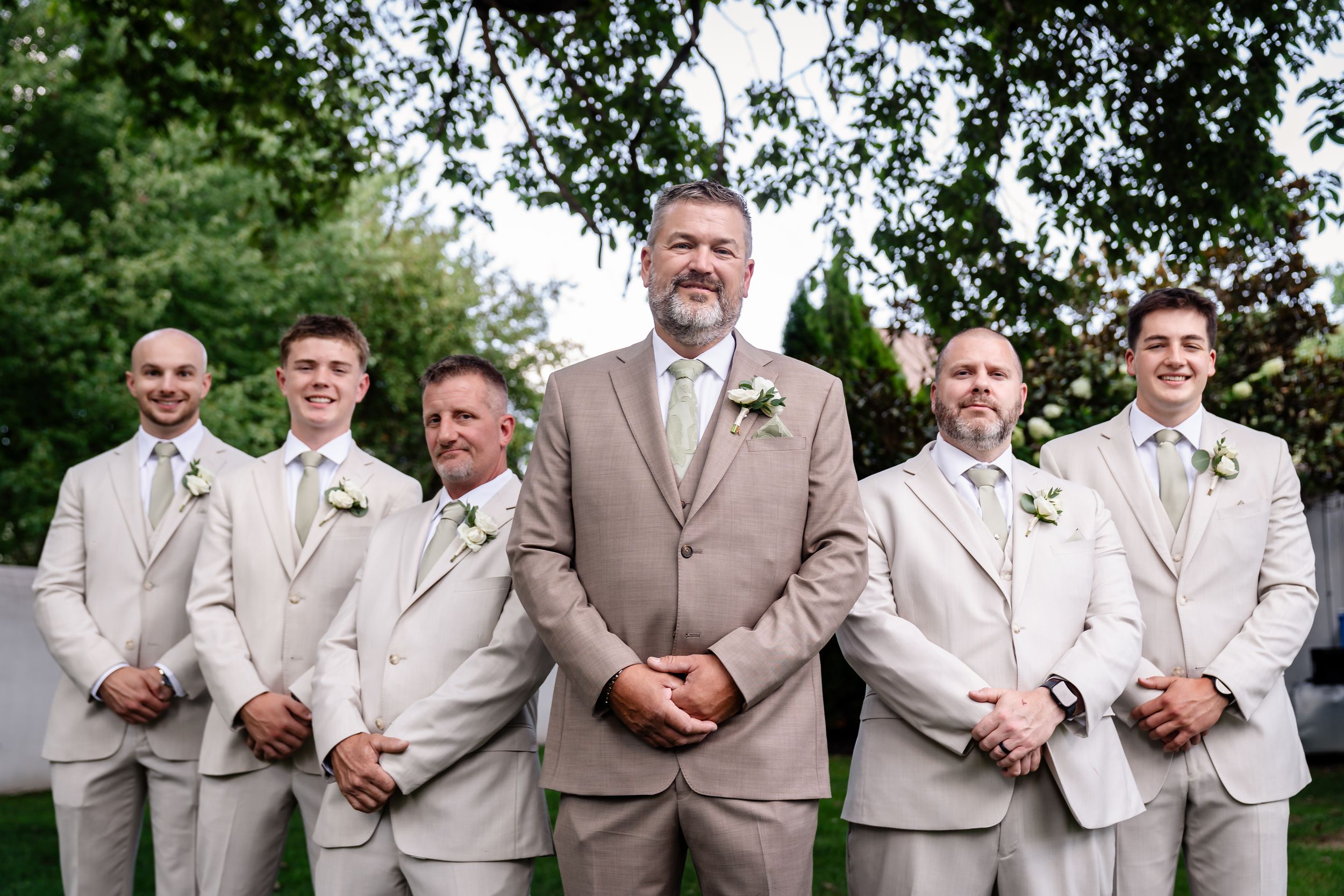 Groom and groomsmen in matching tan suits with white boutonnieres posing outdoors at a wedding ceremony.