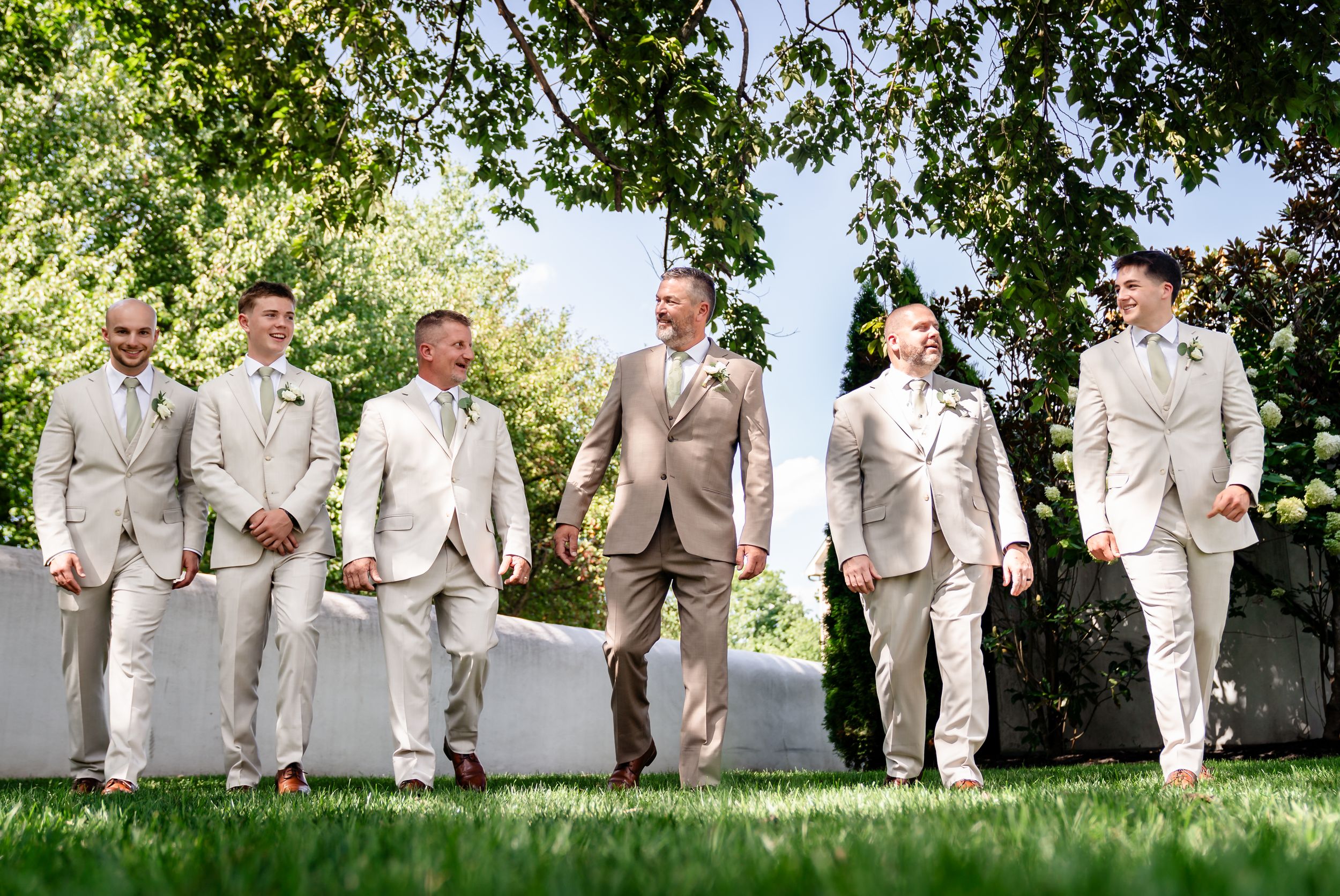 Six groomsmen in matching tan suits walk outdoors on a sunny day at a wedding celebration.