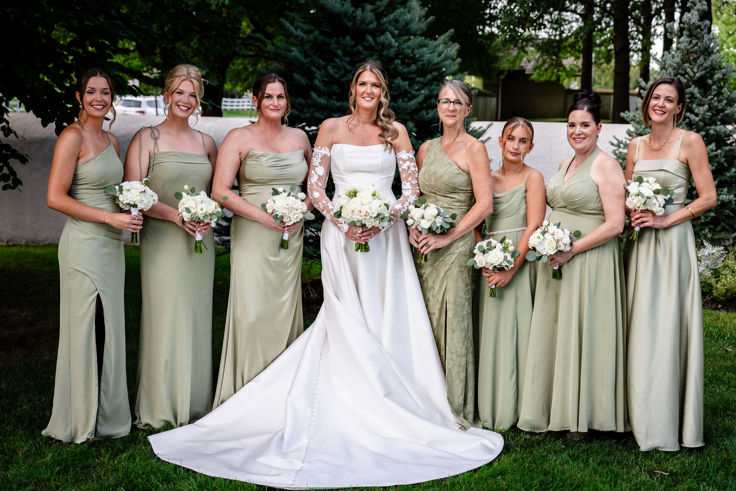 Bride in white gown with bridesmaids in sage green dresses holding white bouquets outdoors at wedding.