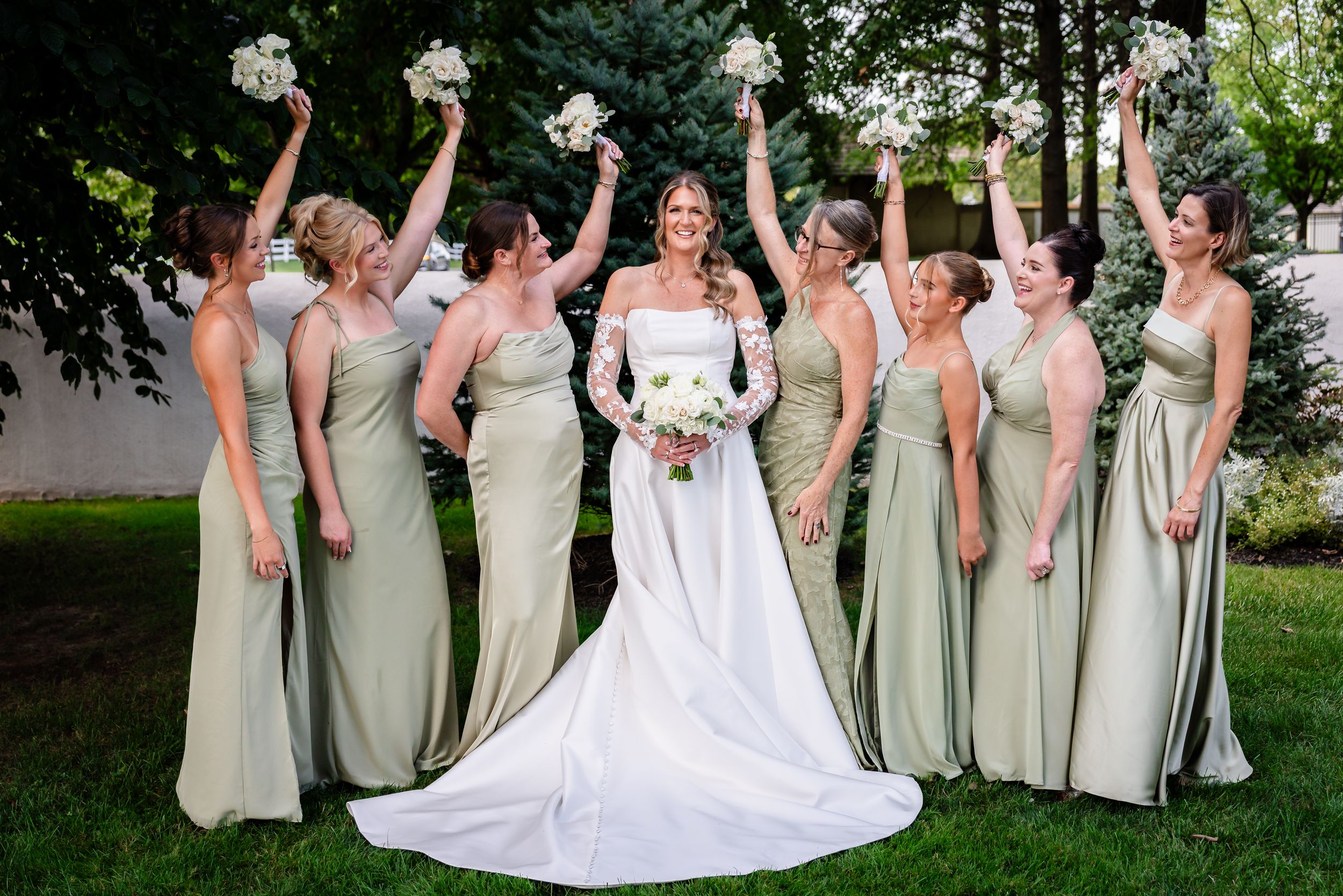 Bride in white gown poses with bridesmaids in sage green dresses raising bouquets outdoors.
