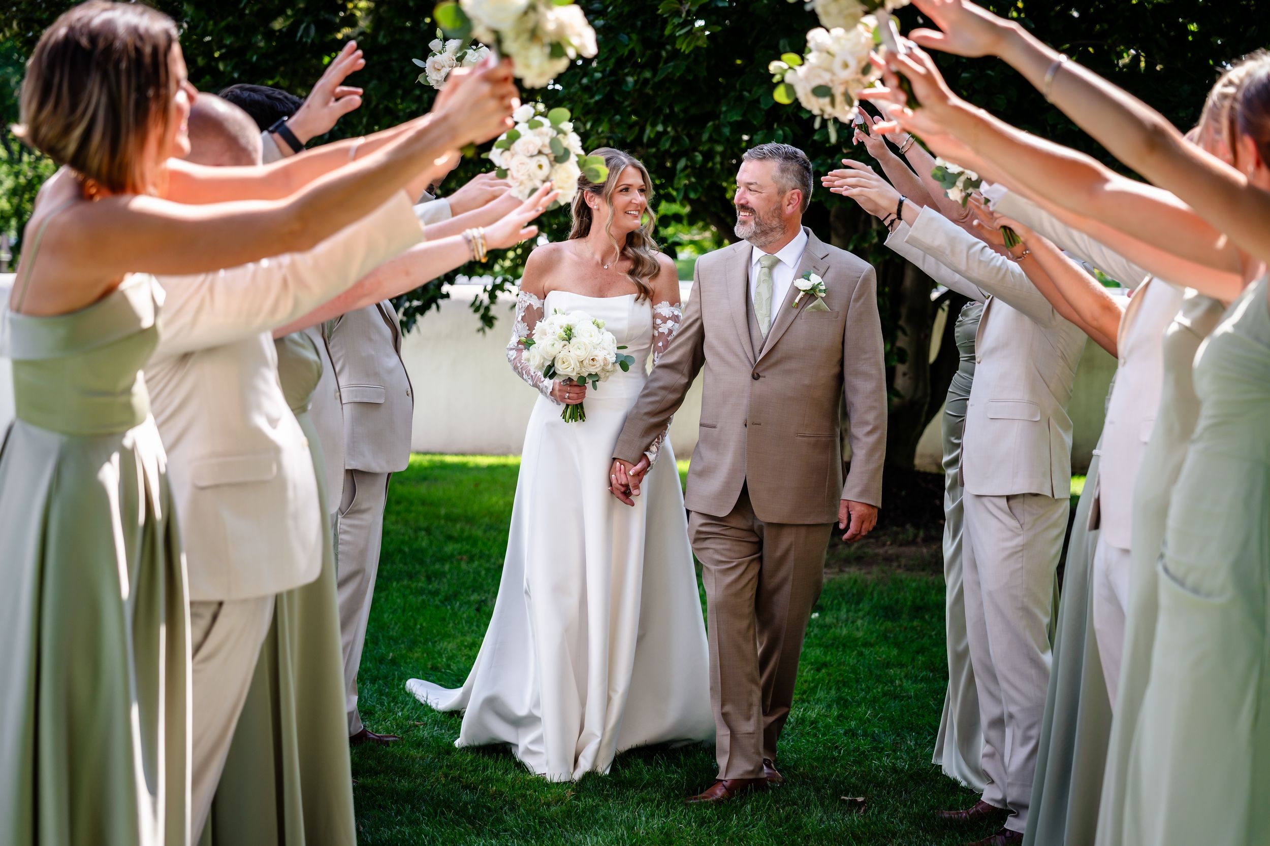 Bride and groom walk through floral arch tunnel created by wedding party in sage green and tan attire outdoors.