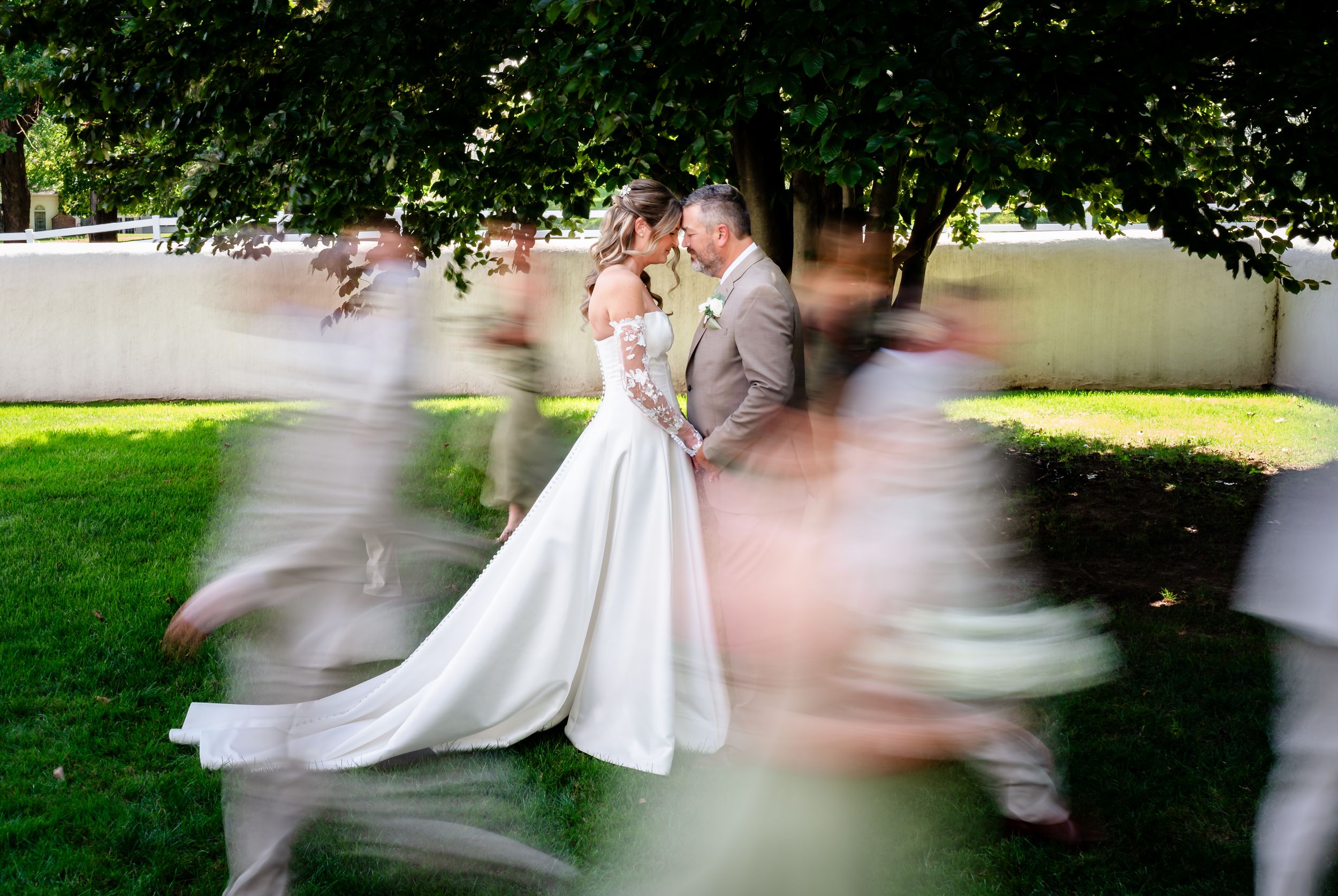 Bride and groom share a romantic kiss while wedding party rushes around them in a blur of motion outdoors.