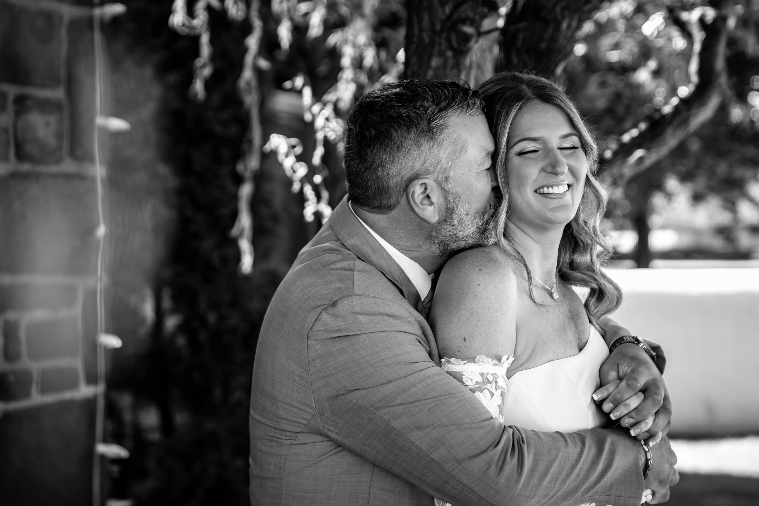 Black and white photo of a groom kissing his bride on the cheek as she smiles joyfully on their wedding day.