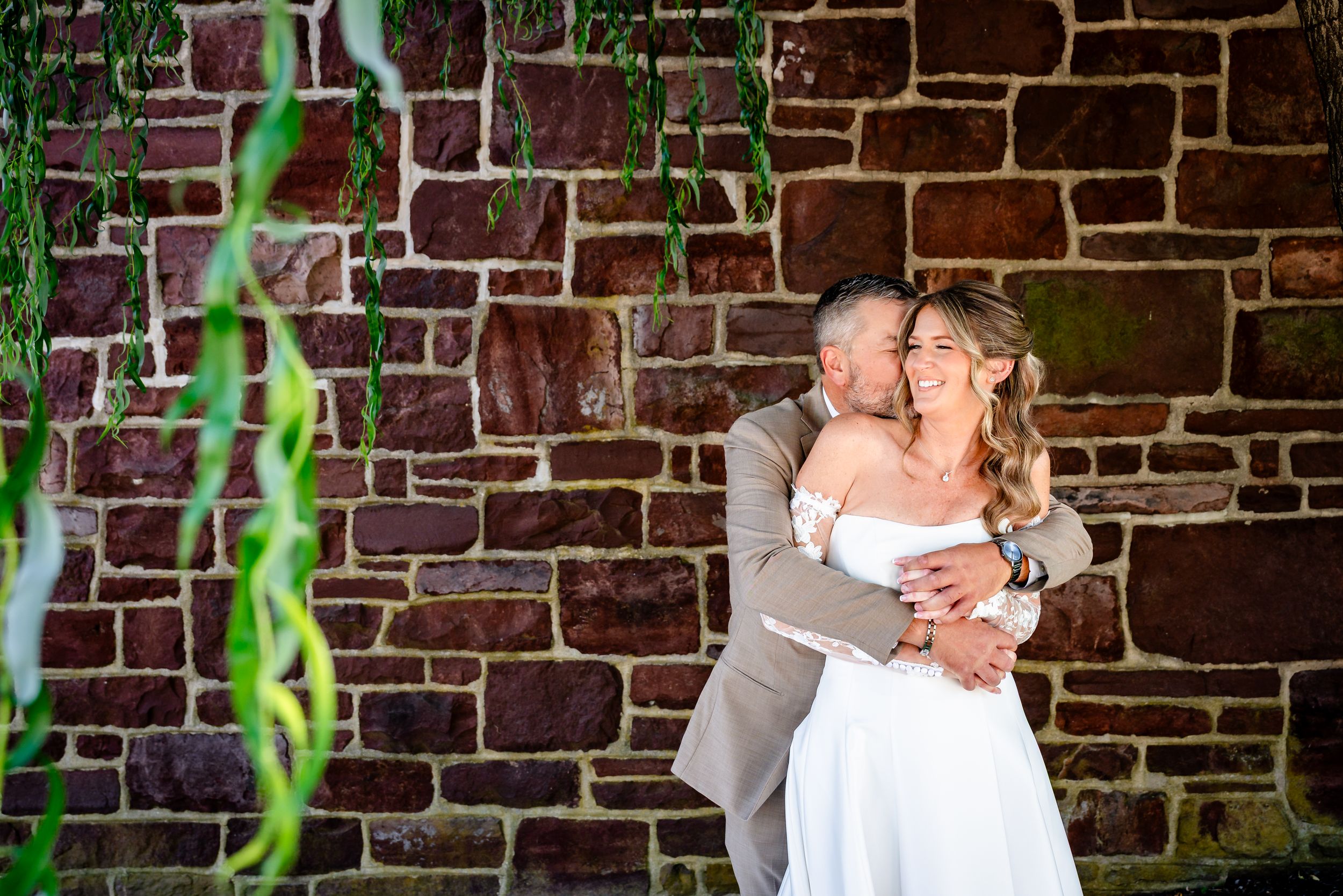 Bride and groom embrace against rustic brick wall with hanging greenery during outdoor wedding portrait.