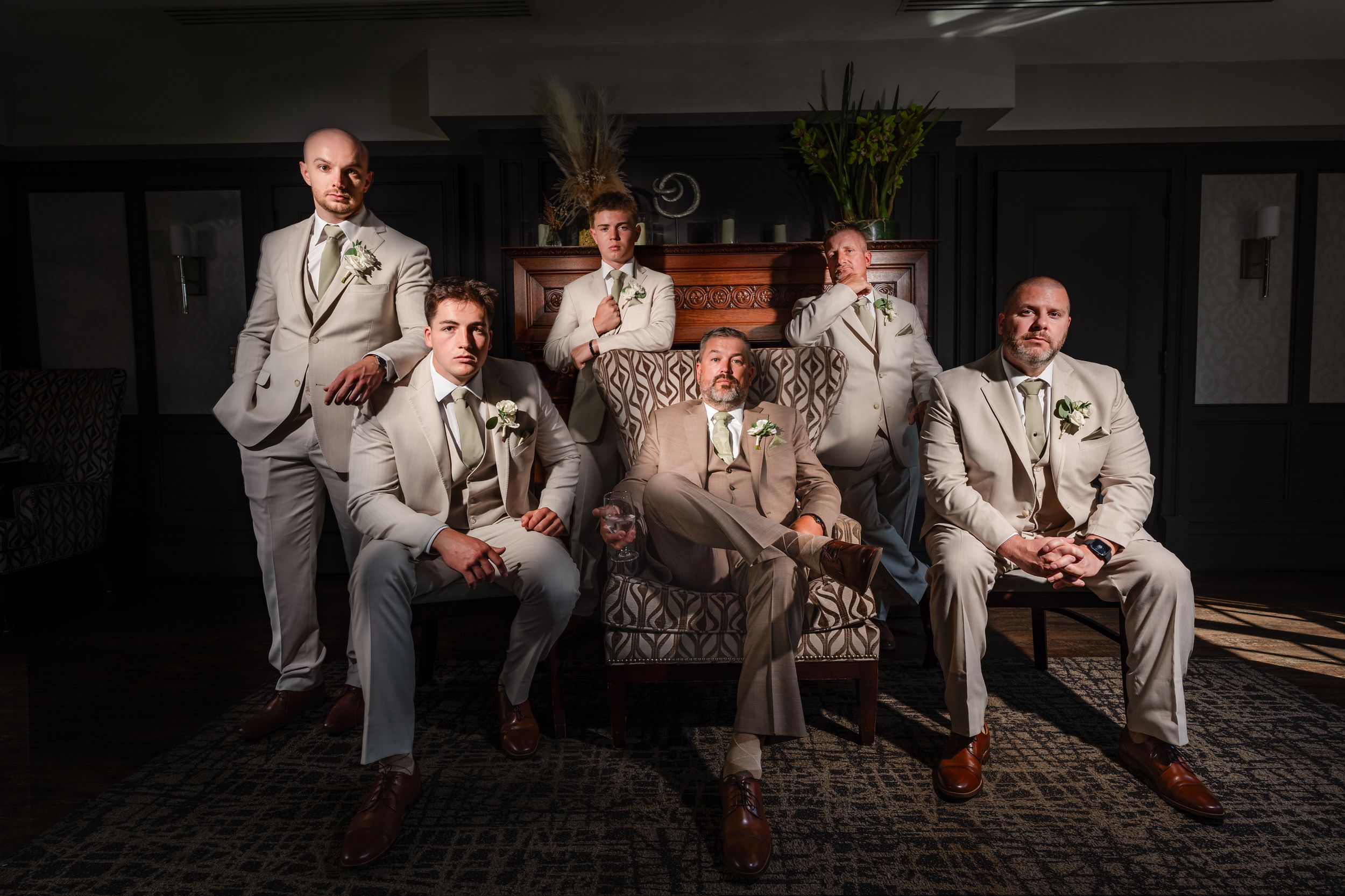 Five groomsmen in matching light gray suits pose dramatically in a dimly lit room for a wedding portrait.