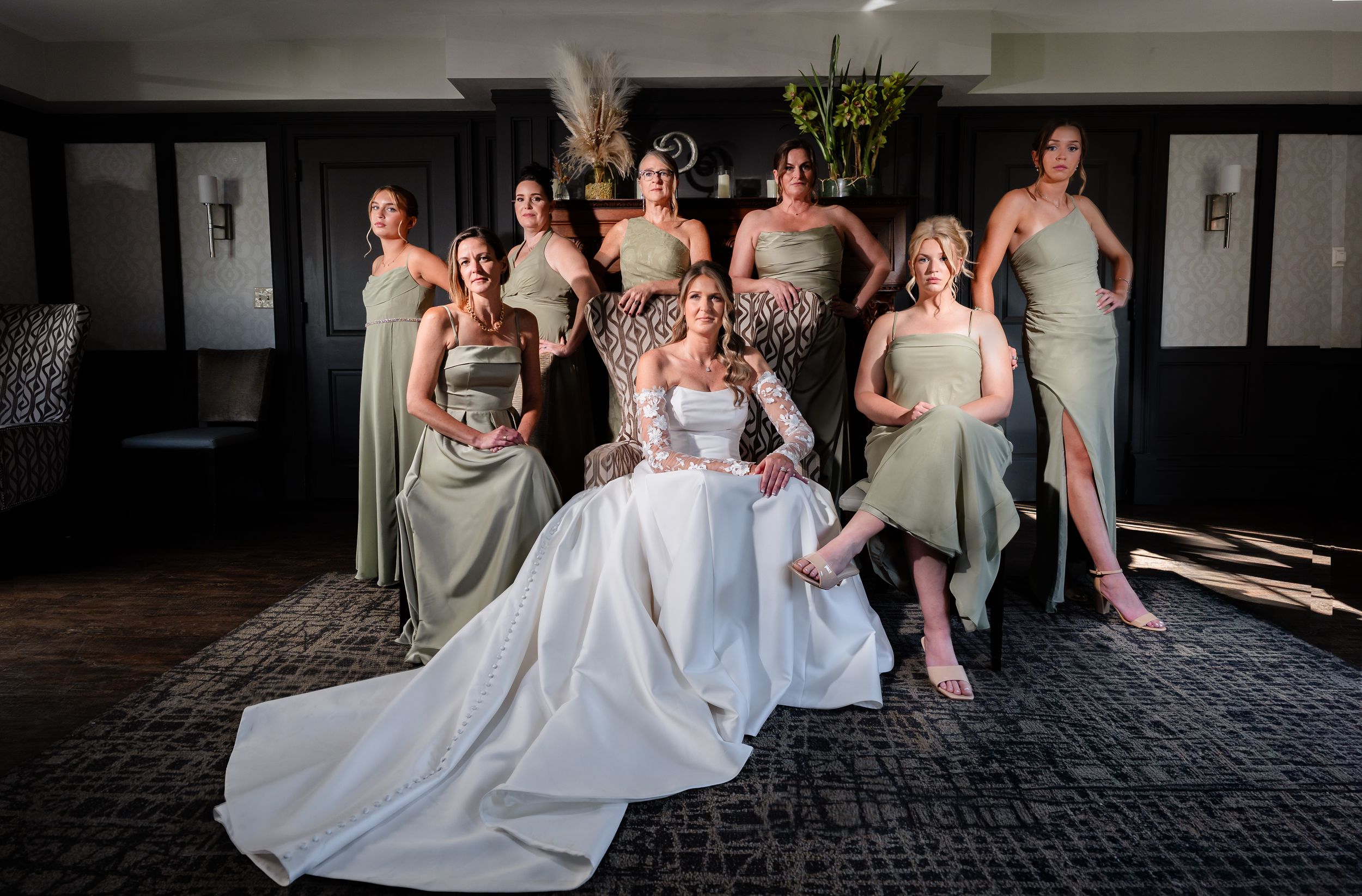 Bride in white gown seated surrounded by eight bridesmaids in sage green dresses in elegant venue.